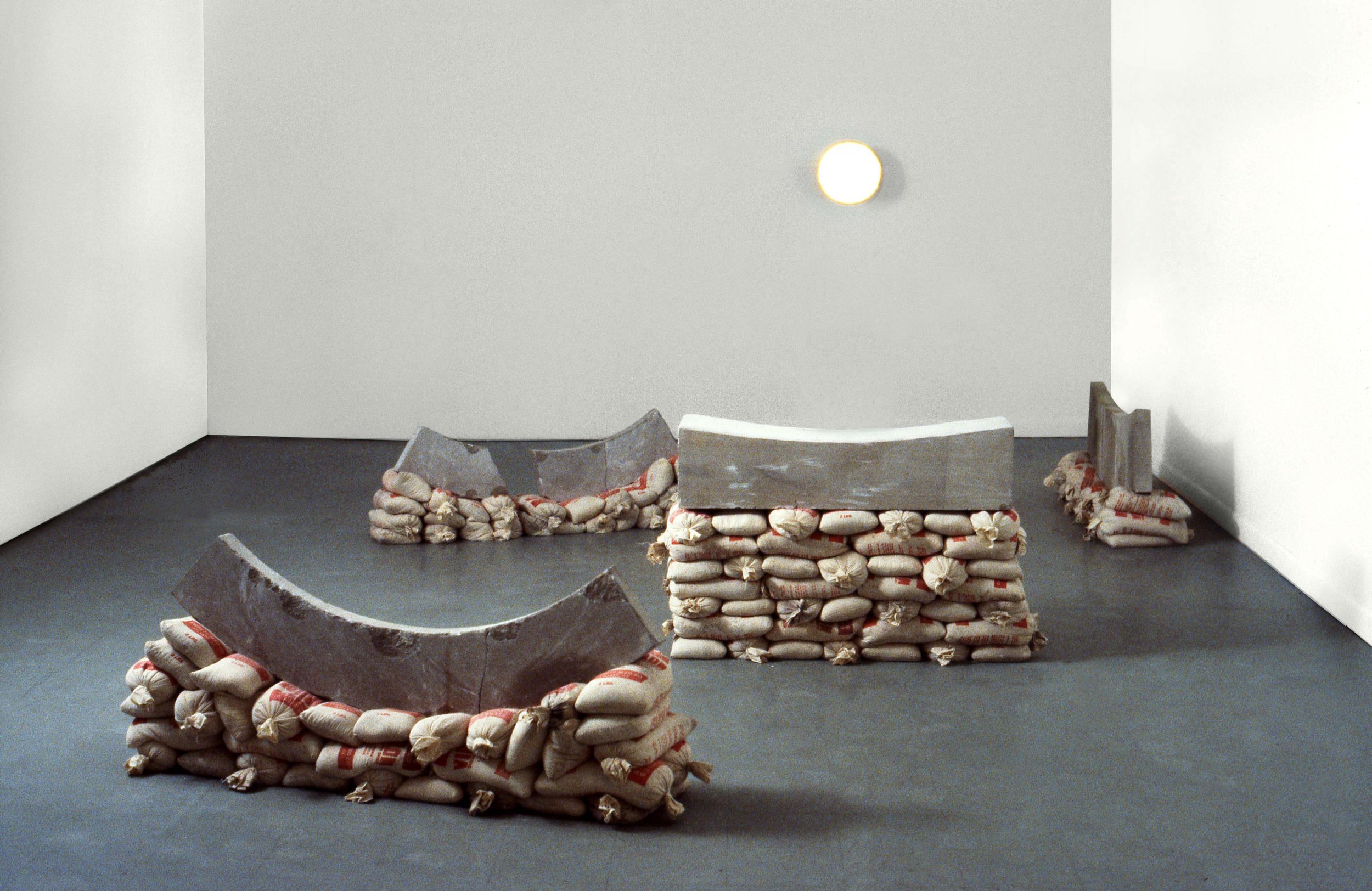 Art installation with concrete curved structures resting on sandbags, arranged in a minimalist room with a gray floor and white walls, illuminated by a single wall light.
