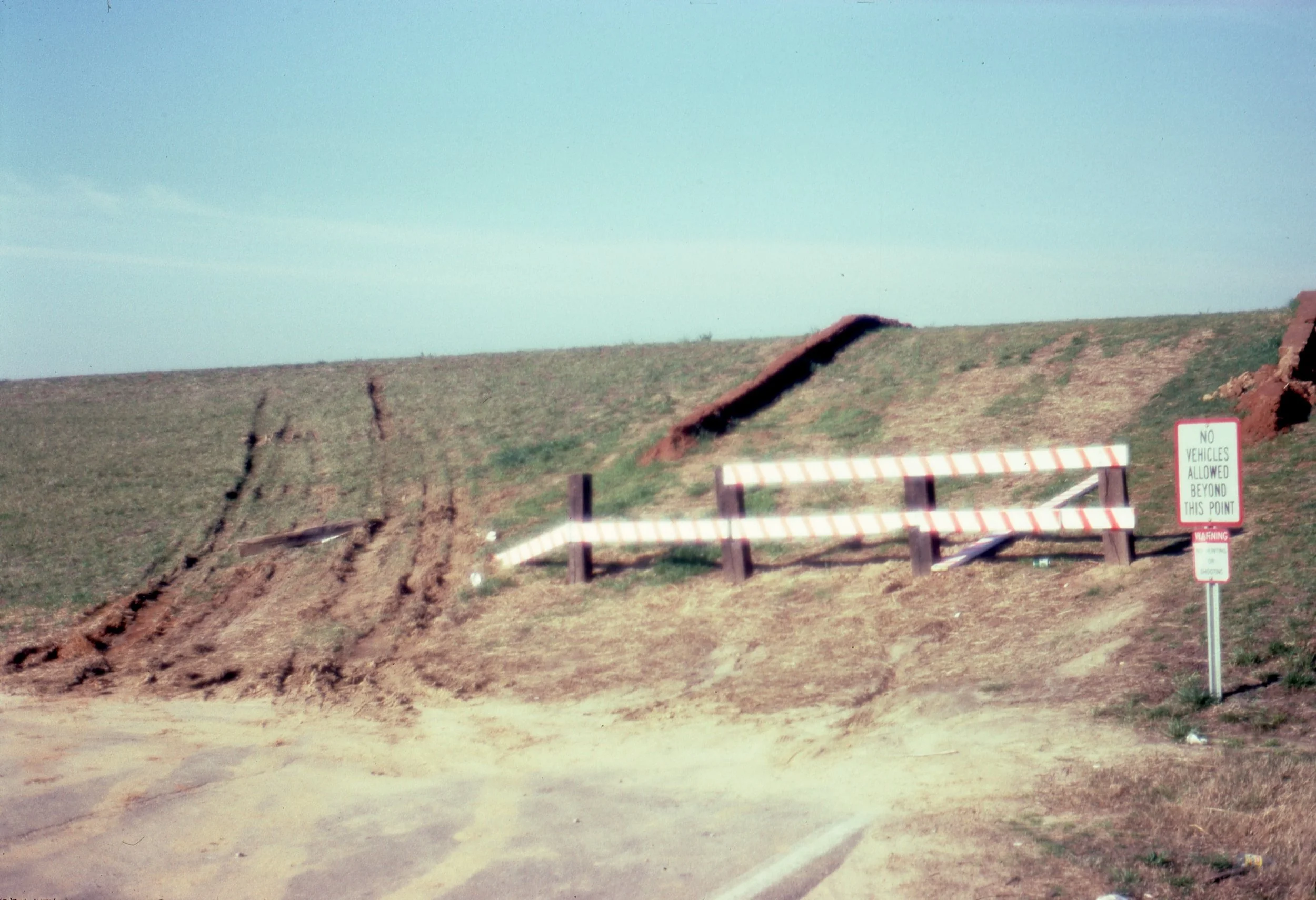 A dirt road blocked by a white and orange striped barrier and a warning sign that says 'No vehicles allowed beyond this point' with visible erosion of the surrounding earth.
