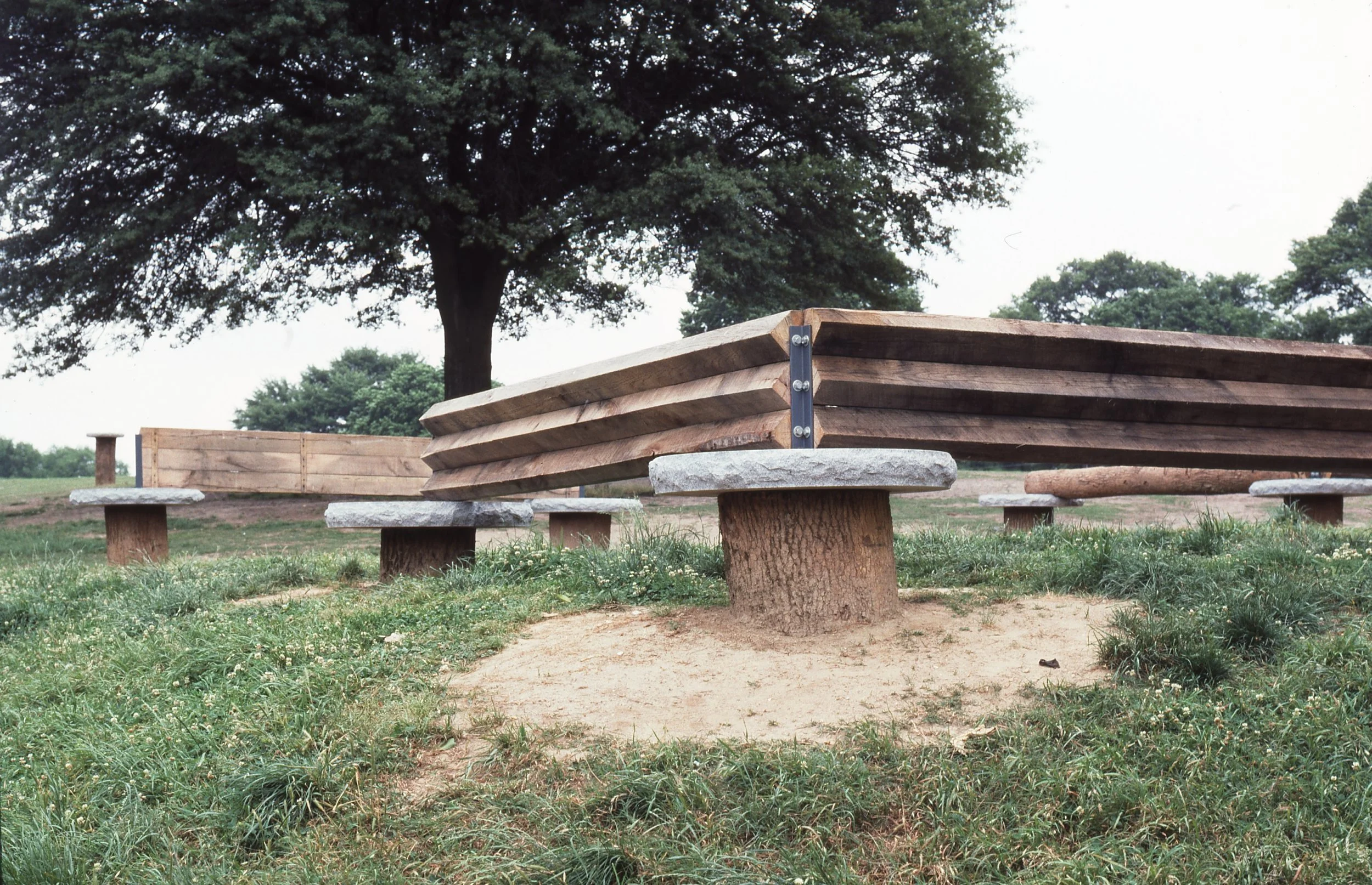 Empty park bench with wooden planks and concrete supports, situated on a grassy area with trees in the background.