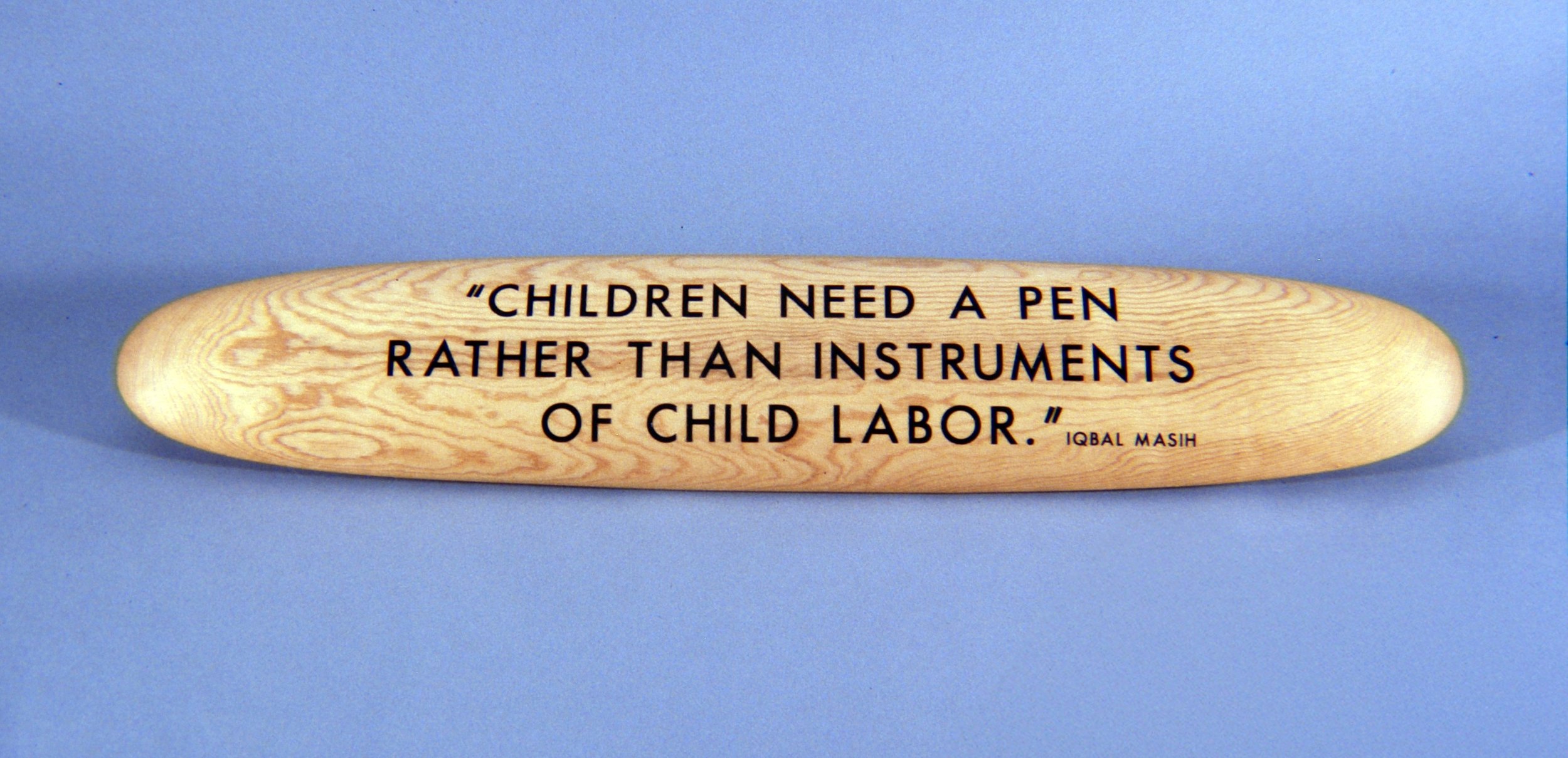 Wooden plaque displaying a quote, reading: 'Children need a pen rather than instruments of child labor.' by Iqbal Masih, set against a blue background.
