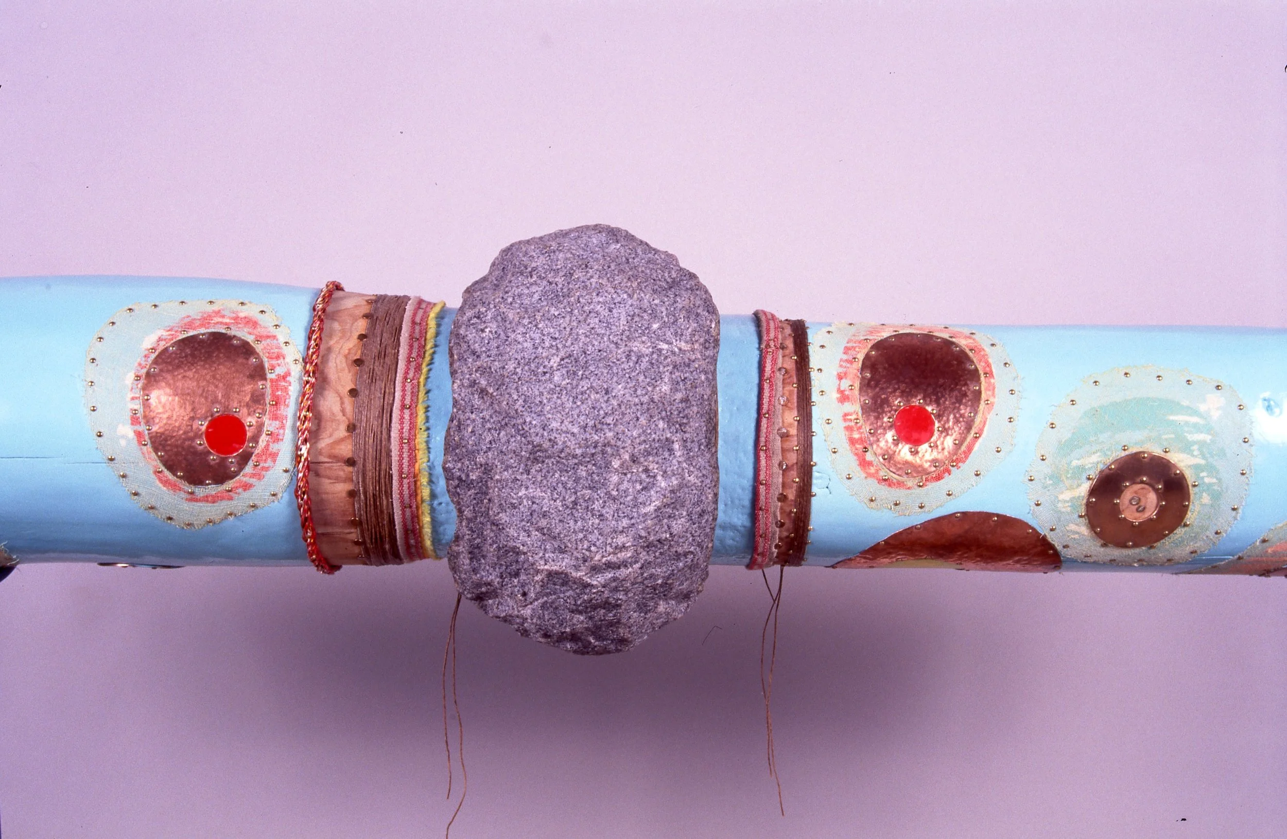 A decorated cylindrical object with colorful patterns and metallic accents, featuring a large gray stone attached to the center.