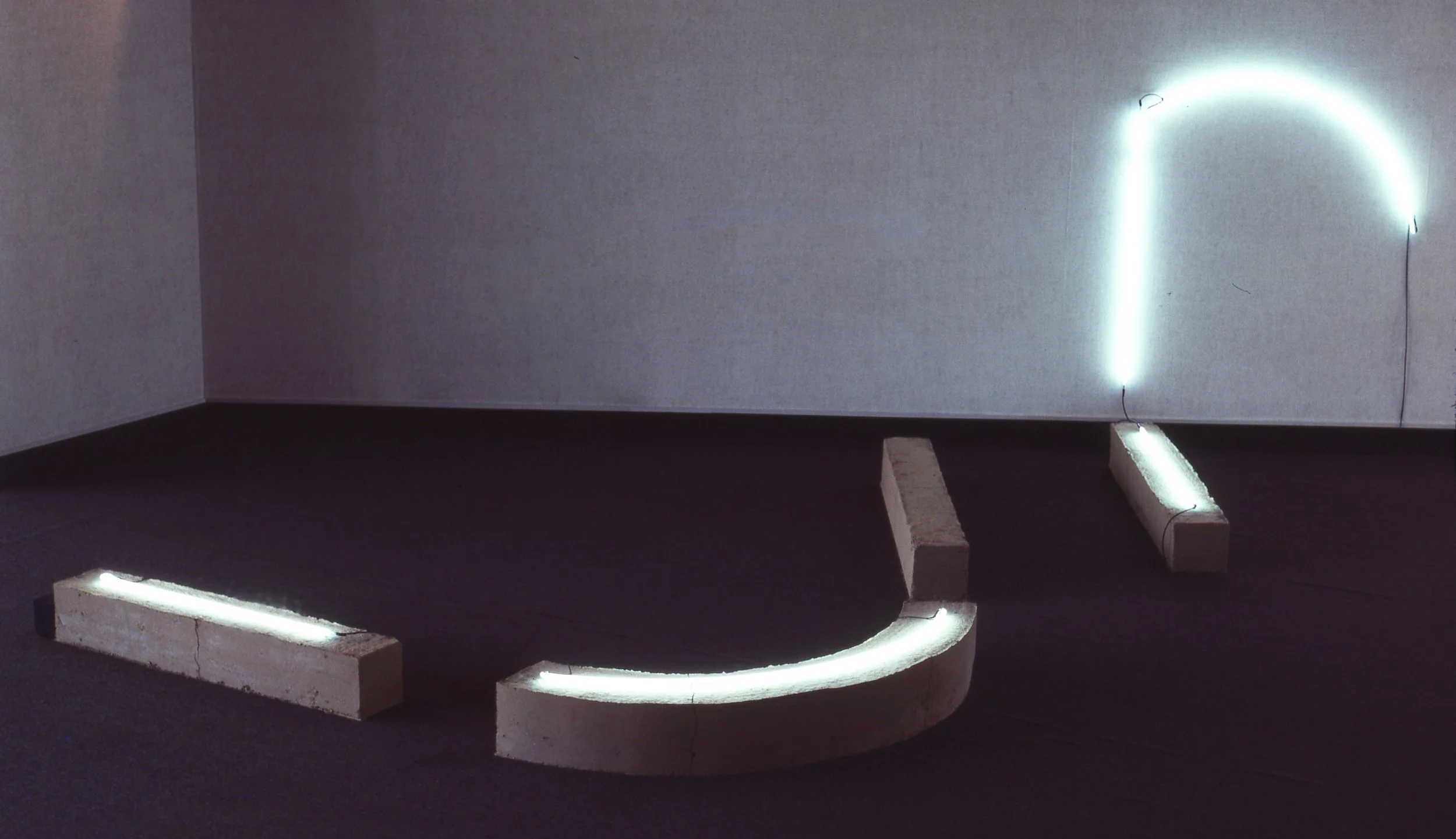 Abstract art installation with concrete blocks and neon lights forming a curved shape on the floor, against a plain wall.
