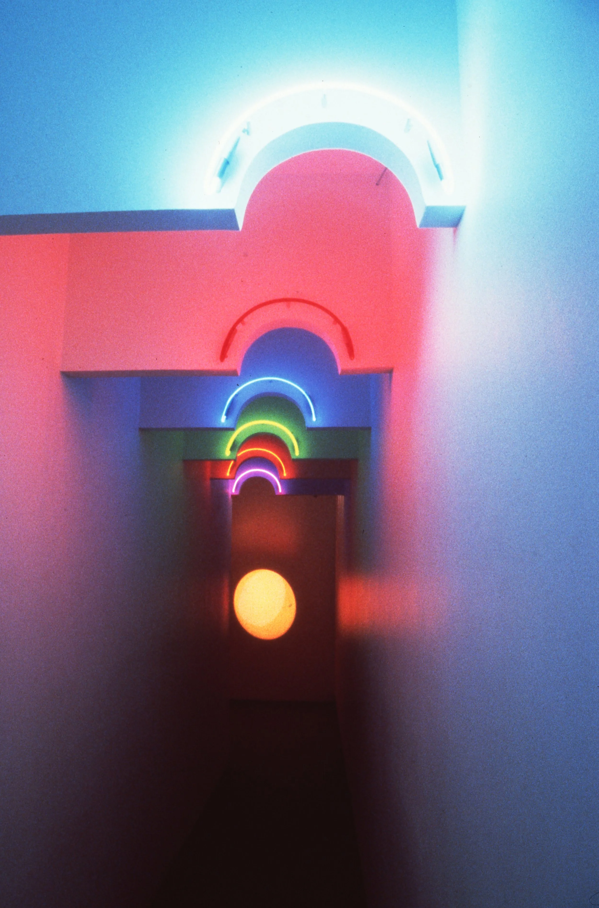 Colorful neon rainbow signs stacked along a hallway ceiling with pastel-colored walls and an oval light fixture at the end.
