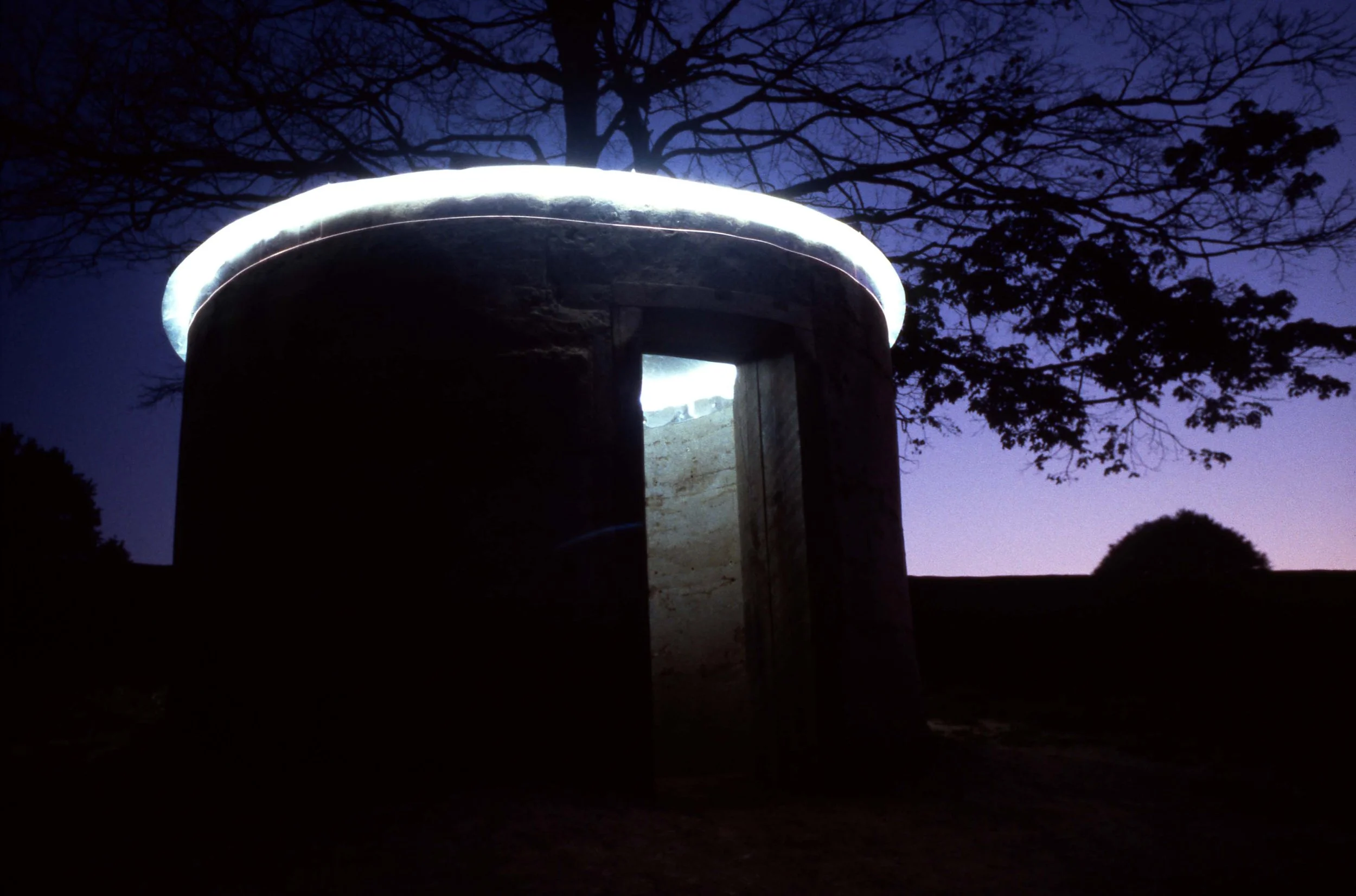 A circular stone structure with an open door is illuminated by a bright white light around the top edge, set against a twilight sky with silhouetted trees in the background.