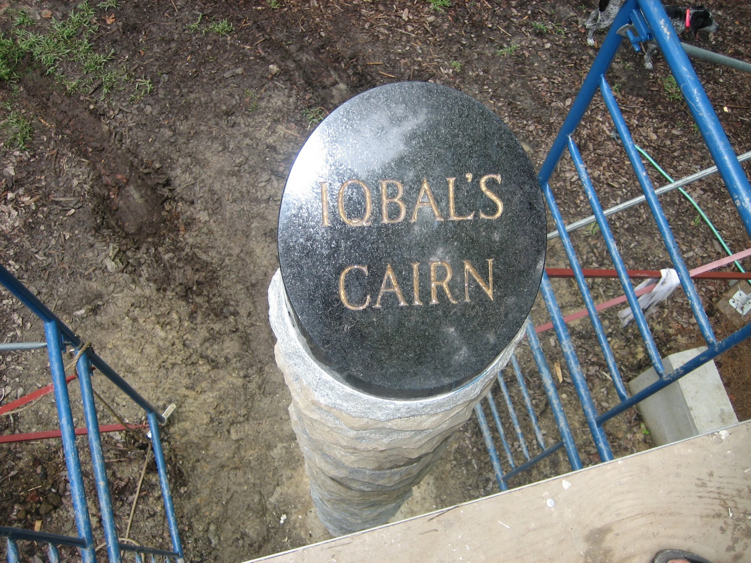 A decorative stone pillar with a black engraved plaque that reads 'IQBAL'S CAIRN'. The pillar is surrounded by a blue metal fence and the ground appears to be dirt or soil.