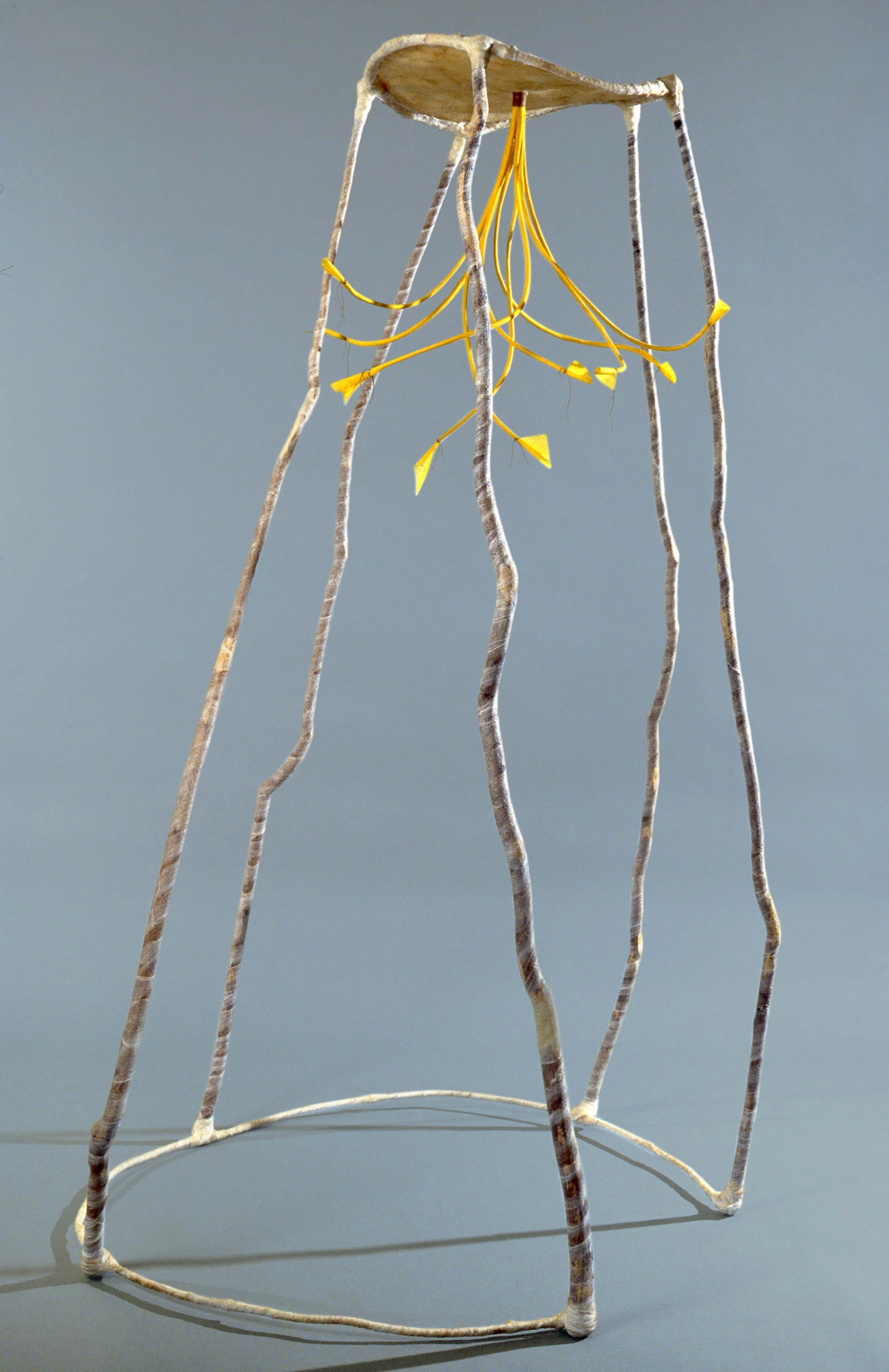 An abstract sculpture consisting of thin, wavy, elongated metallic rods forming a tall, open framework. Yellow wires hang from the top, draping down and connecting to the rods.