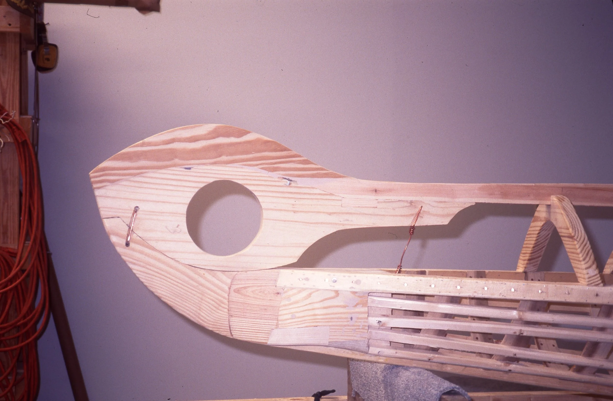 Wooden structure in progress, possibly part of a boat or a decorative piece, with cutouts and slats, mounted against a plain light purple wall.
