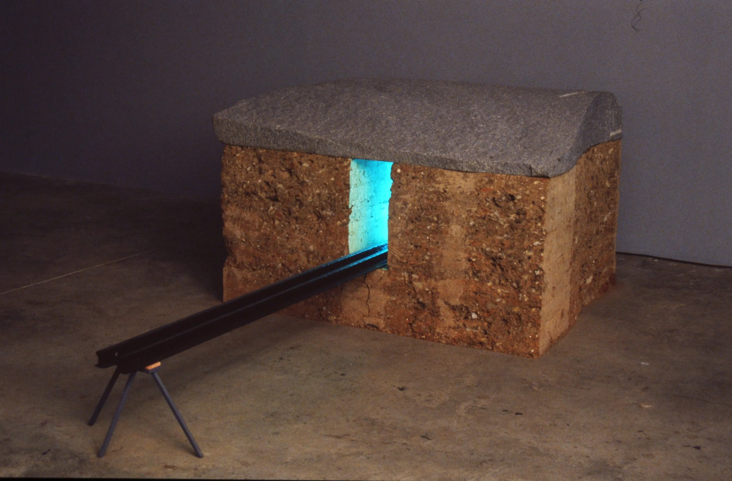 An art installation built of a brick-like structure with a blue light shining through a narrow opening, and a long black rod or tube extending from the opening, supported by a small tripod stand.