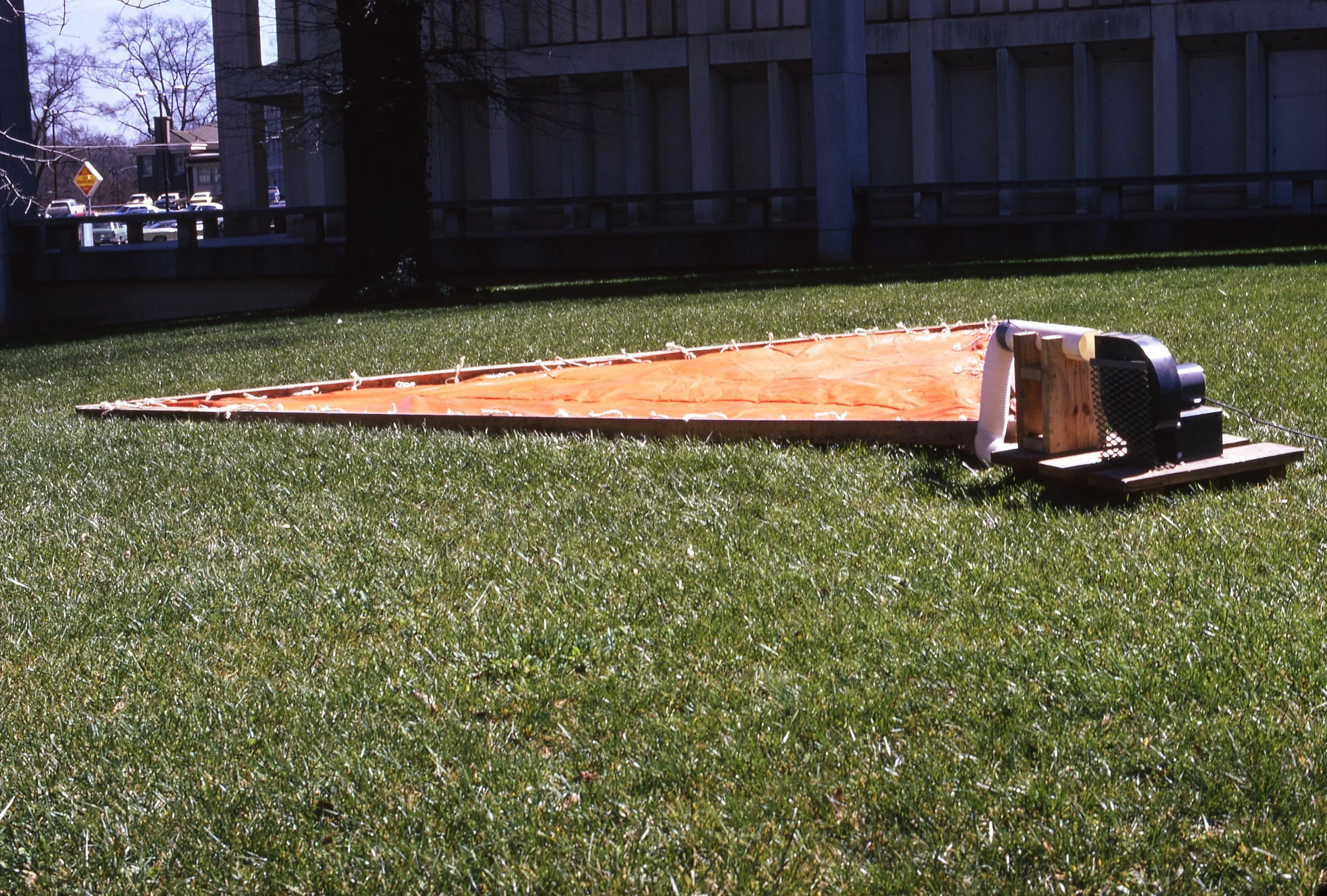 Unassembled large orange hot air balloon basket frame with a fan and other equipment on a grassy lawn, with a fence and trees in the background.