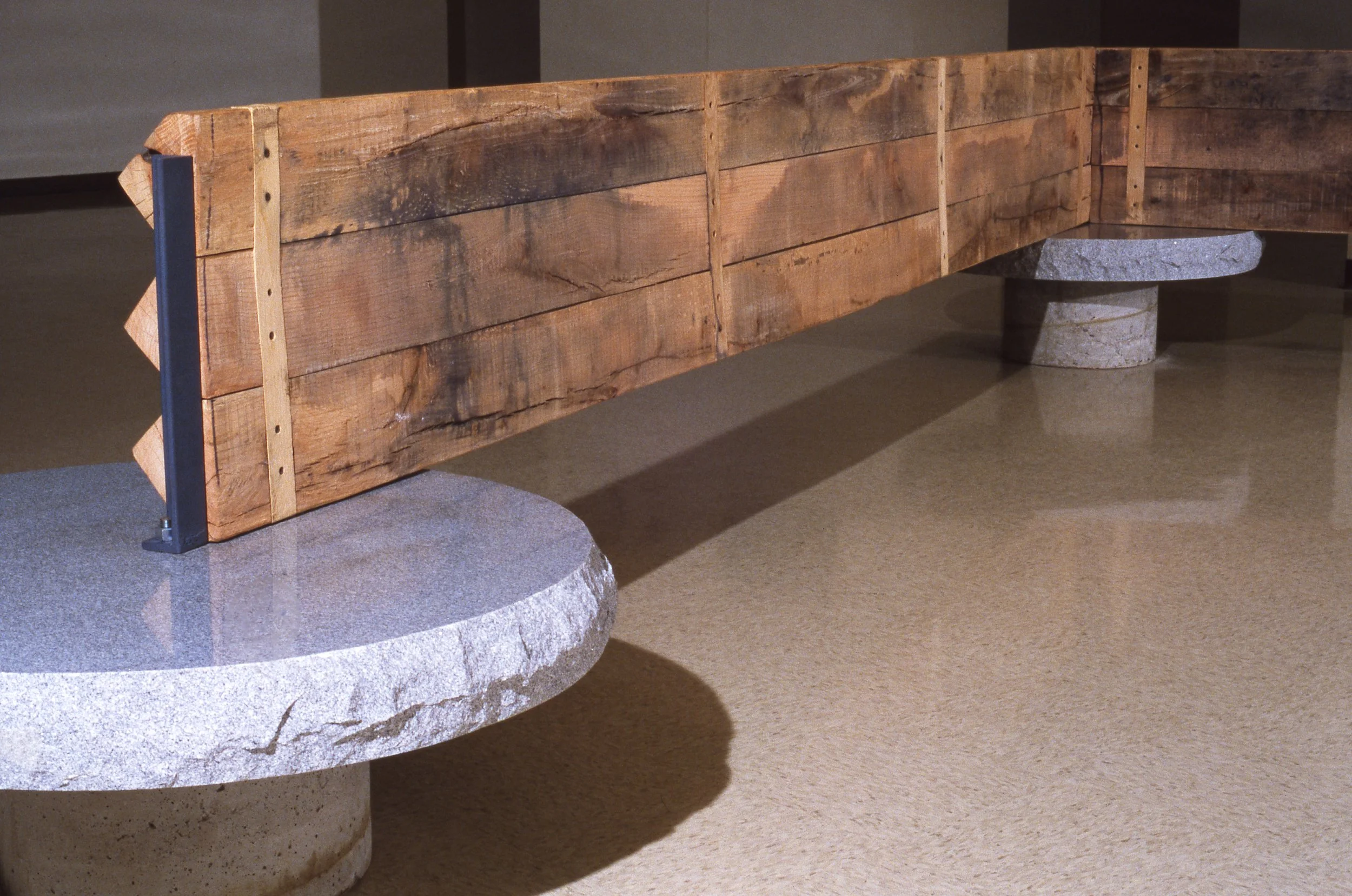 A wooden bench with a rustic appearance, supported by two cylindrical stone bases, is displayed indoors.