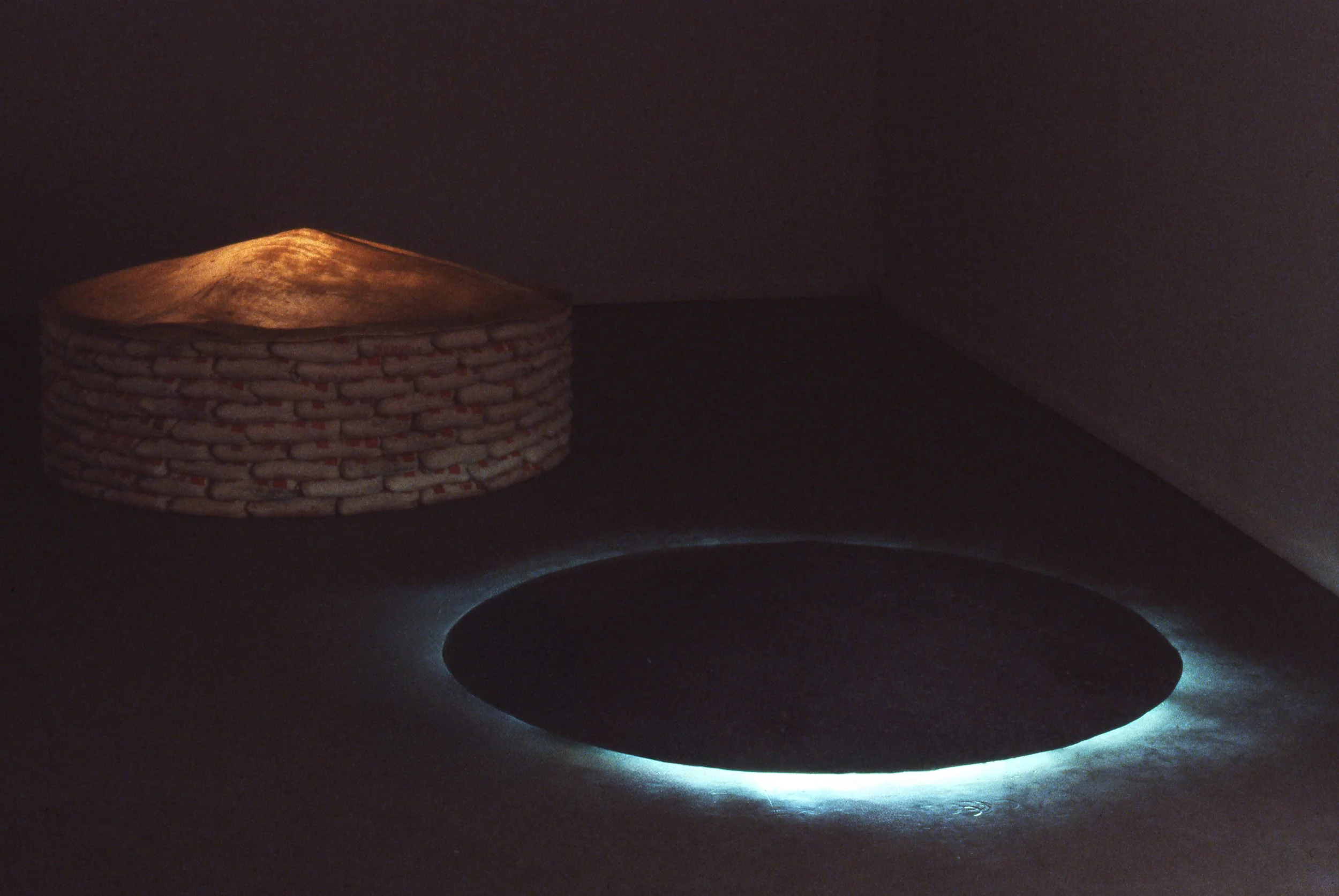 A circular hole in the floor with light shining through, and a small, rounded, brick structure nearby topped with a glowing, cone-shaped light.