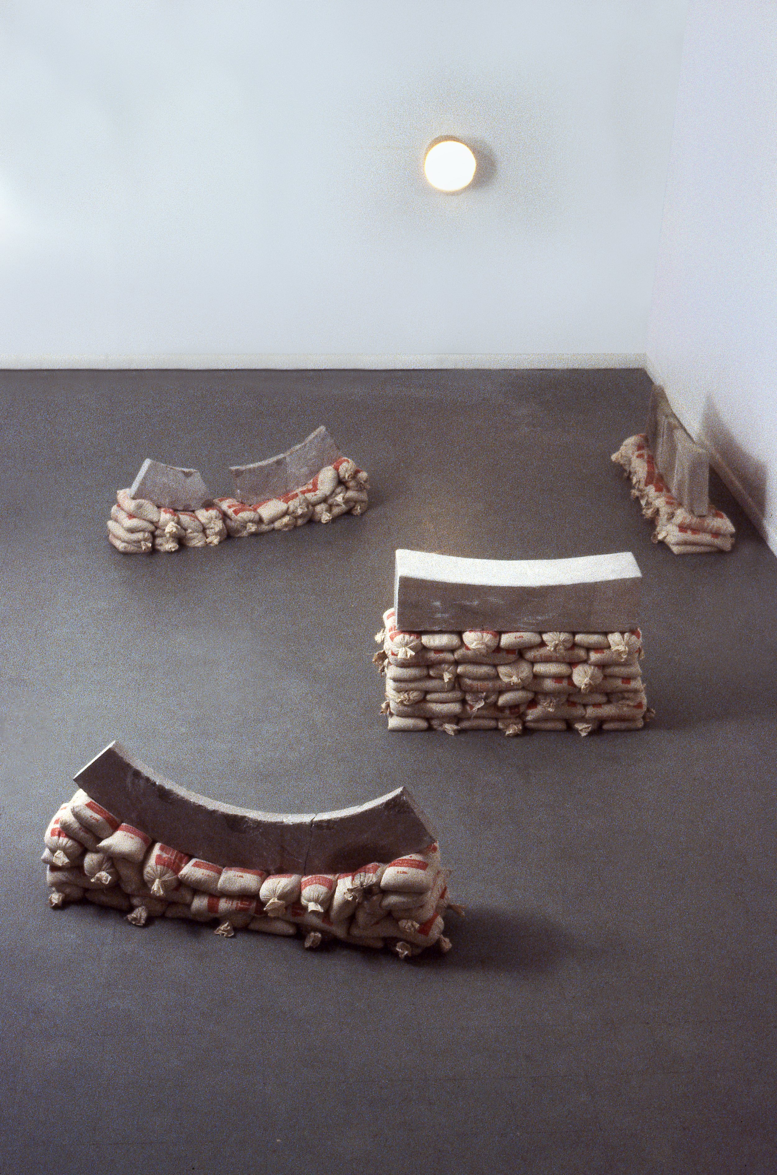 Abstract art installation with curved and rectangular forms made of layered bags or fabrics, arranged on a dark floor in a room with white walls and a ceiling light.