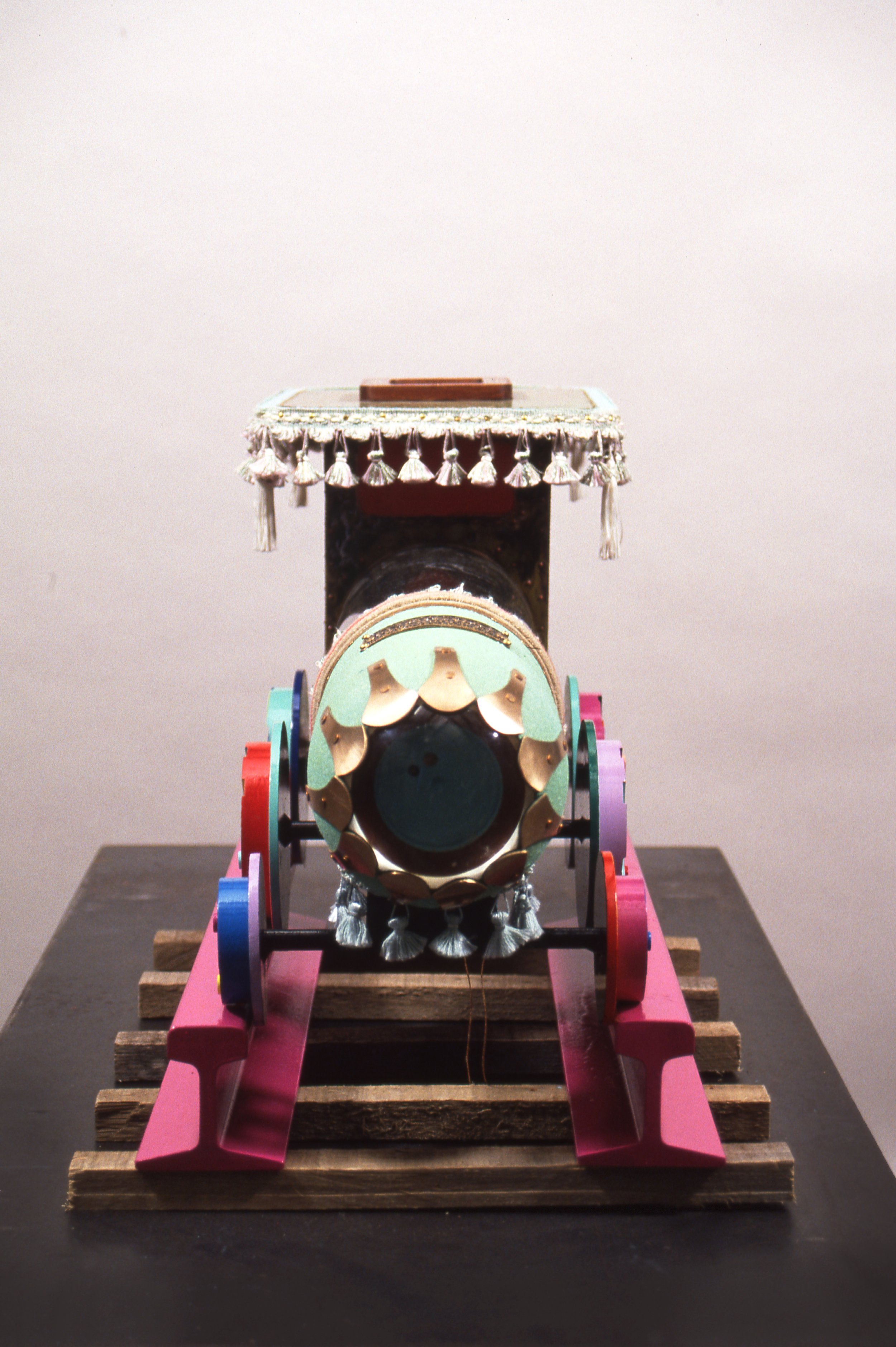 A small decorated wooden cart with a box on top, positioned on a display stand against a plain background.