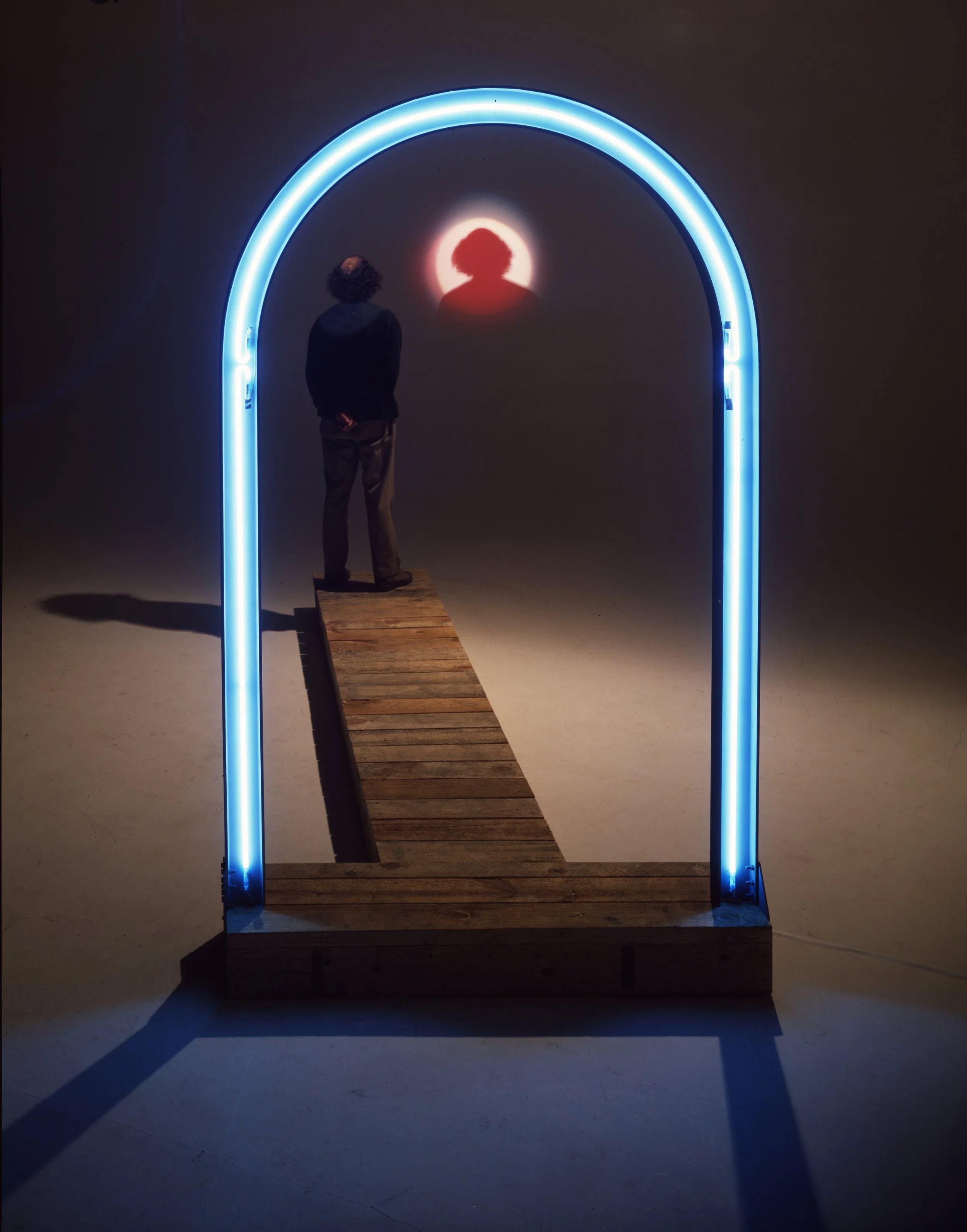 A person standing on a wooden platform in front of a wall with a glowing silhouette of a woman with an afro hairstyle, illuminated by a neon arch.