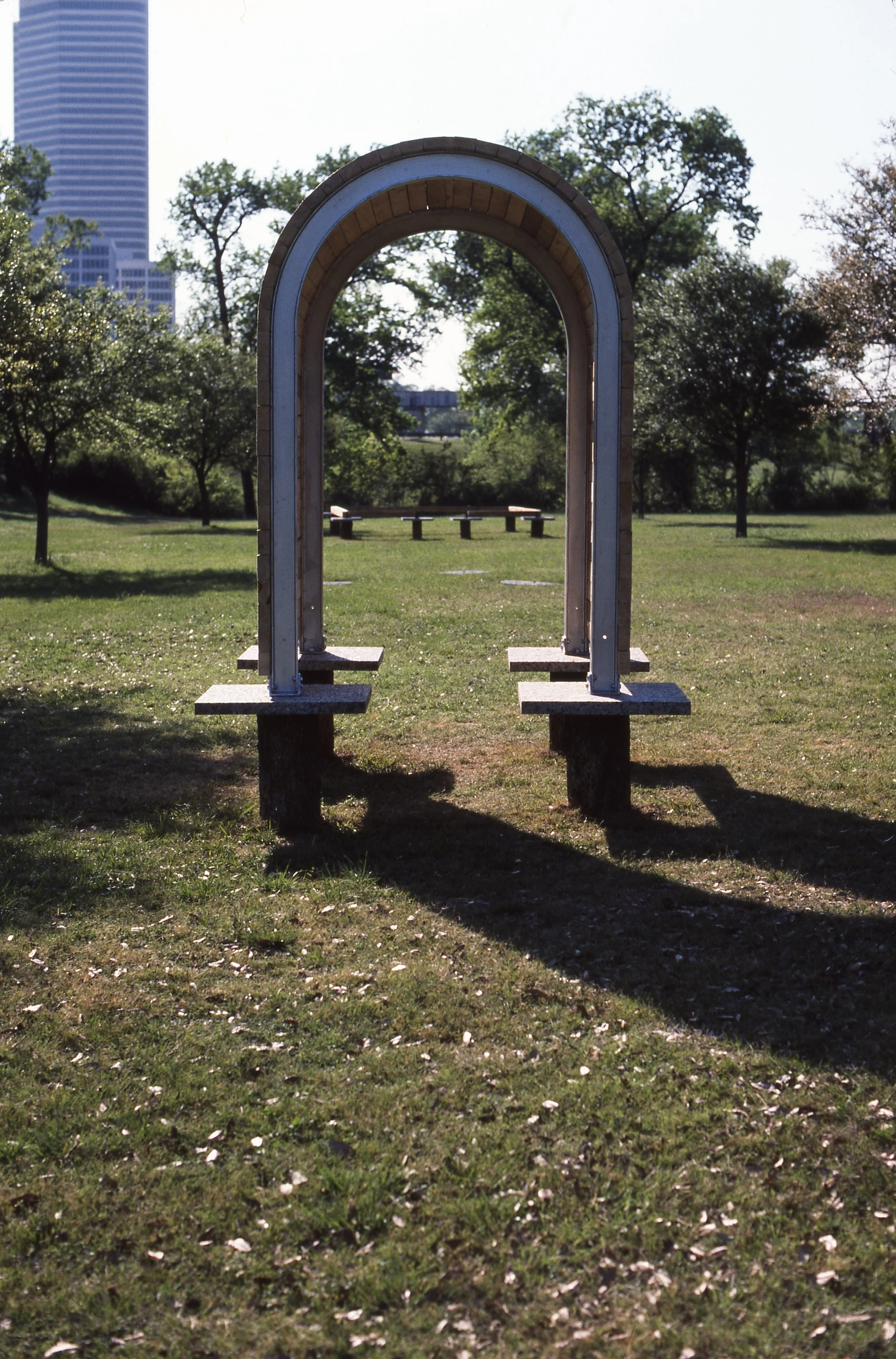 A park with a metal archway and benches, with trees and a tall building in the background.