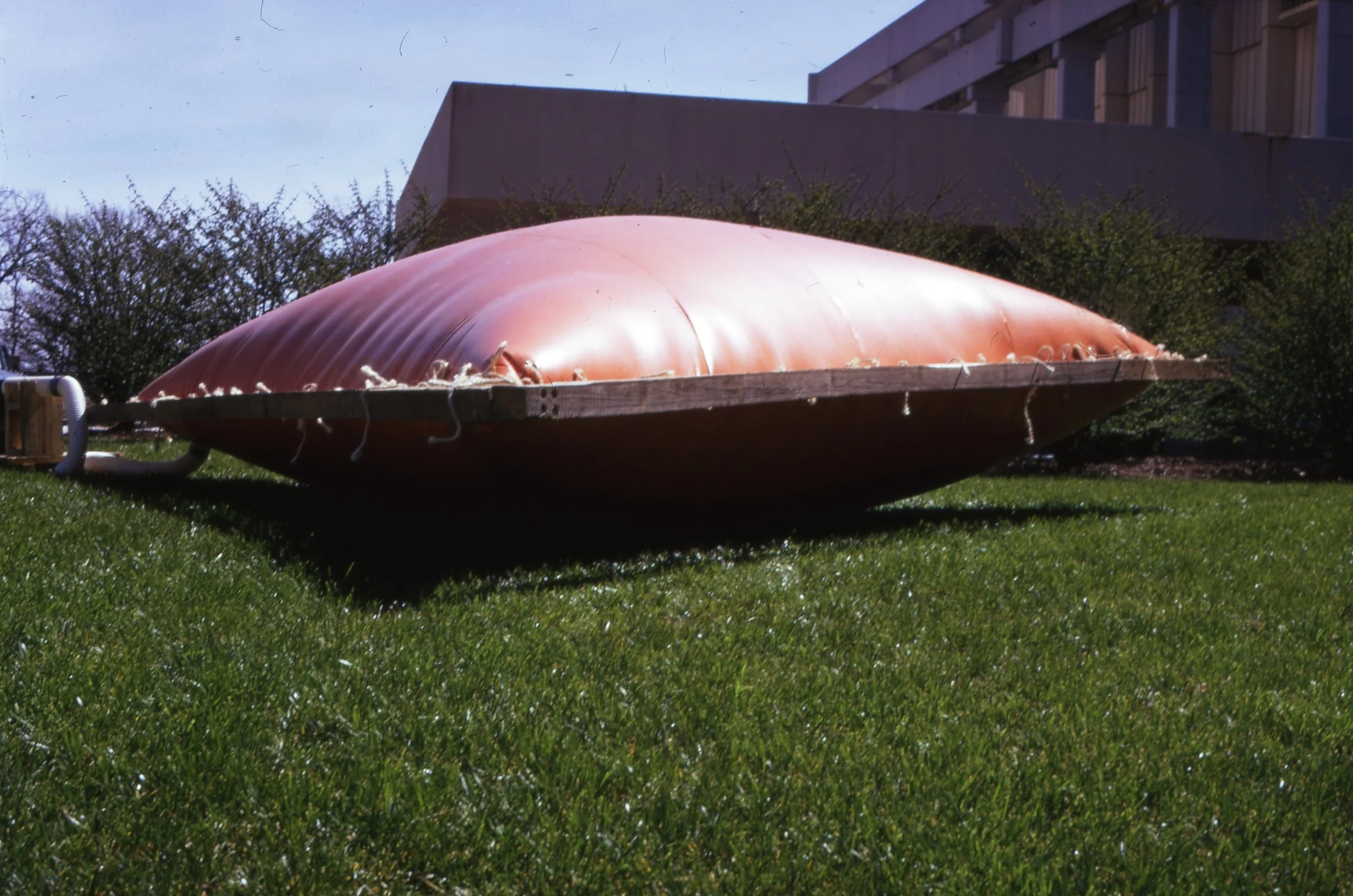 Large red inflatable boat with a wooden base lying on green grass, lightly tethered with ropes, with bushes and a building in the background under a clear sky.