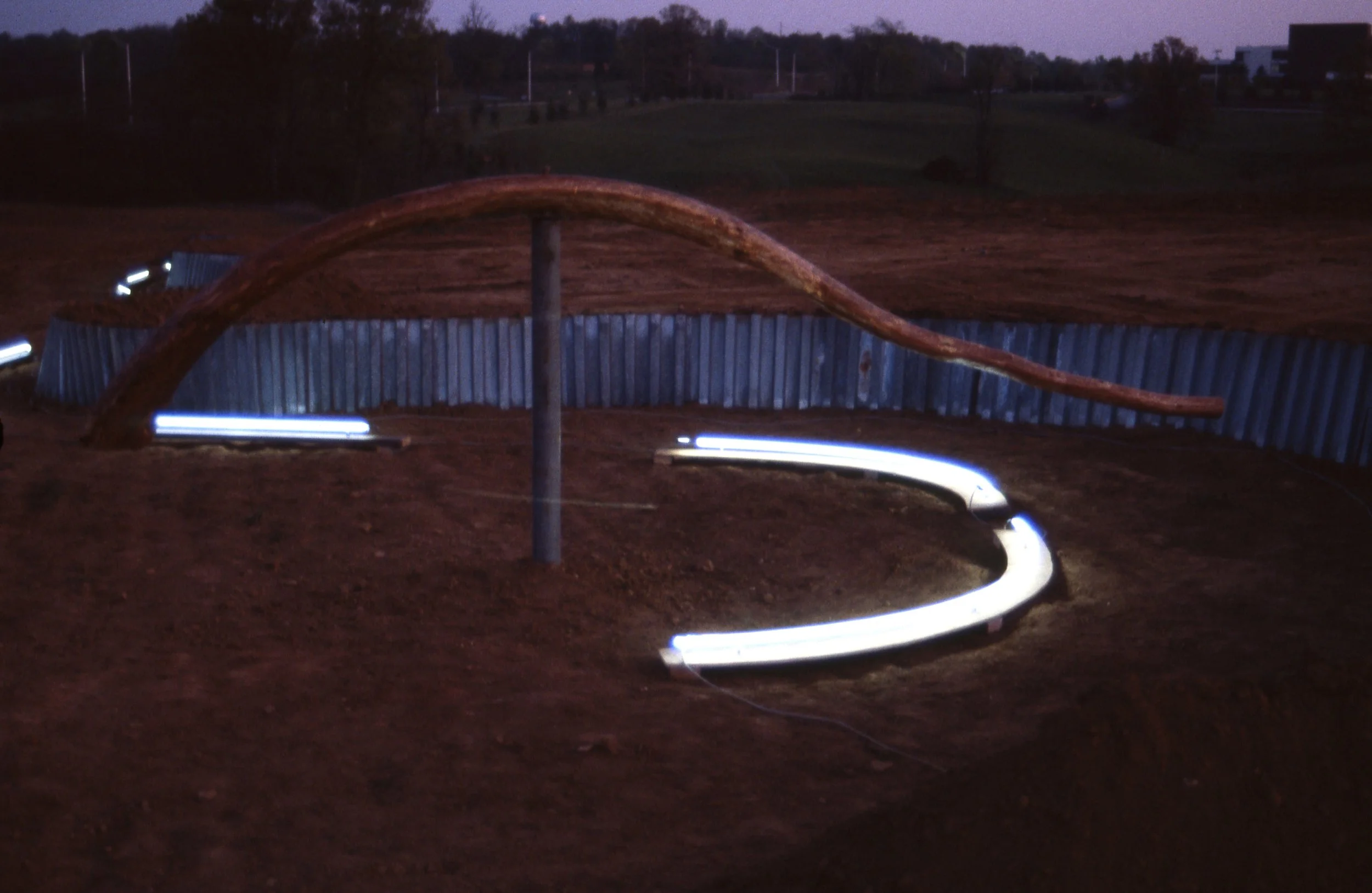 An outdoor children’s playground at dusk featuring a curved slide with illuminated steps and a tall, curved wooden climbing structure, surrounded by soil and a metal fence.