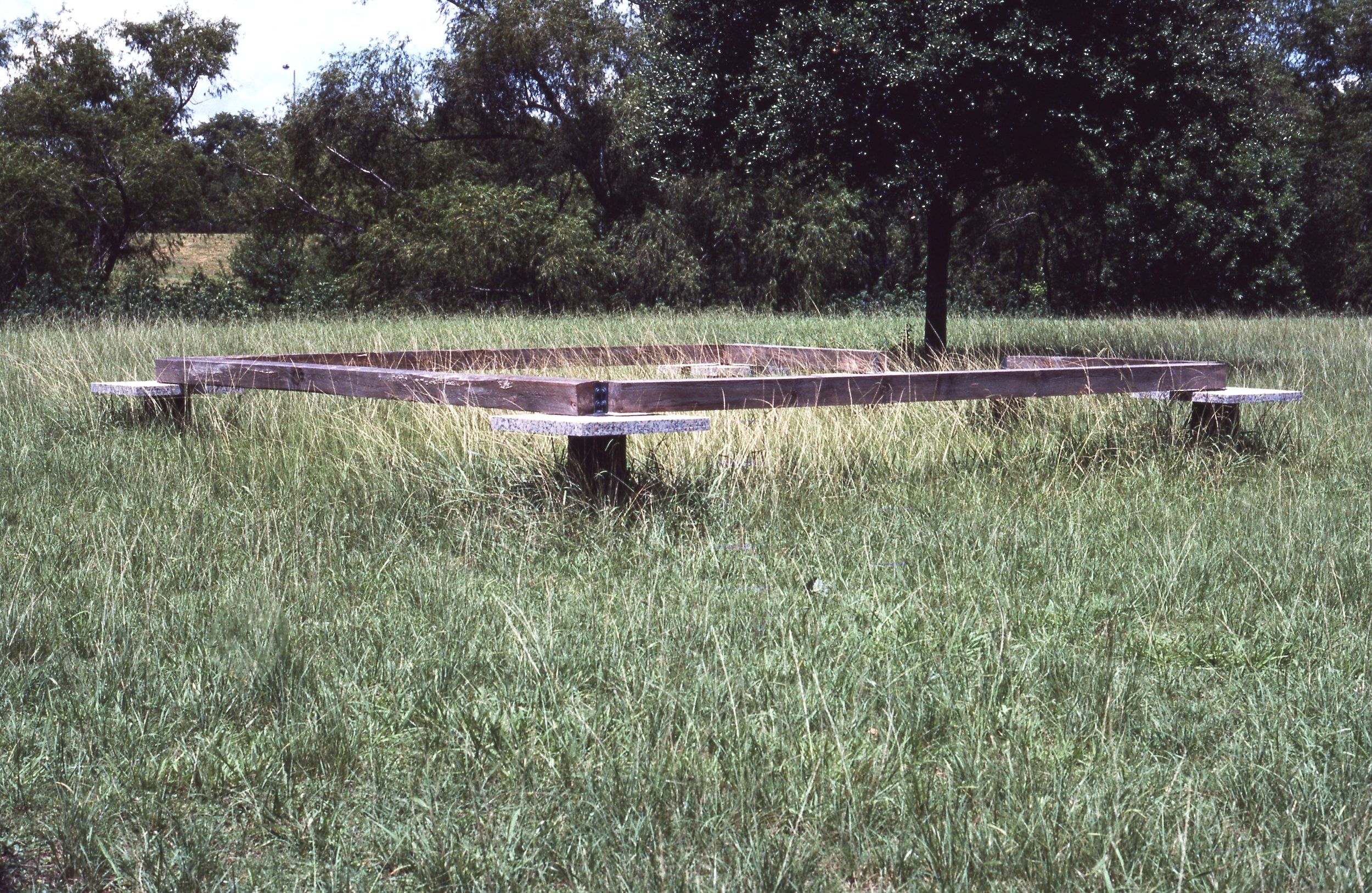 Empty wooden-frame structure on the grass in an open field with trees in the background.