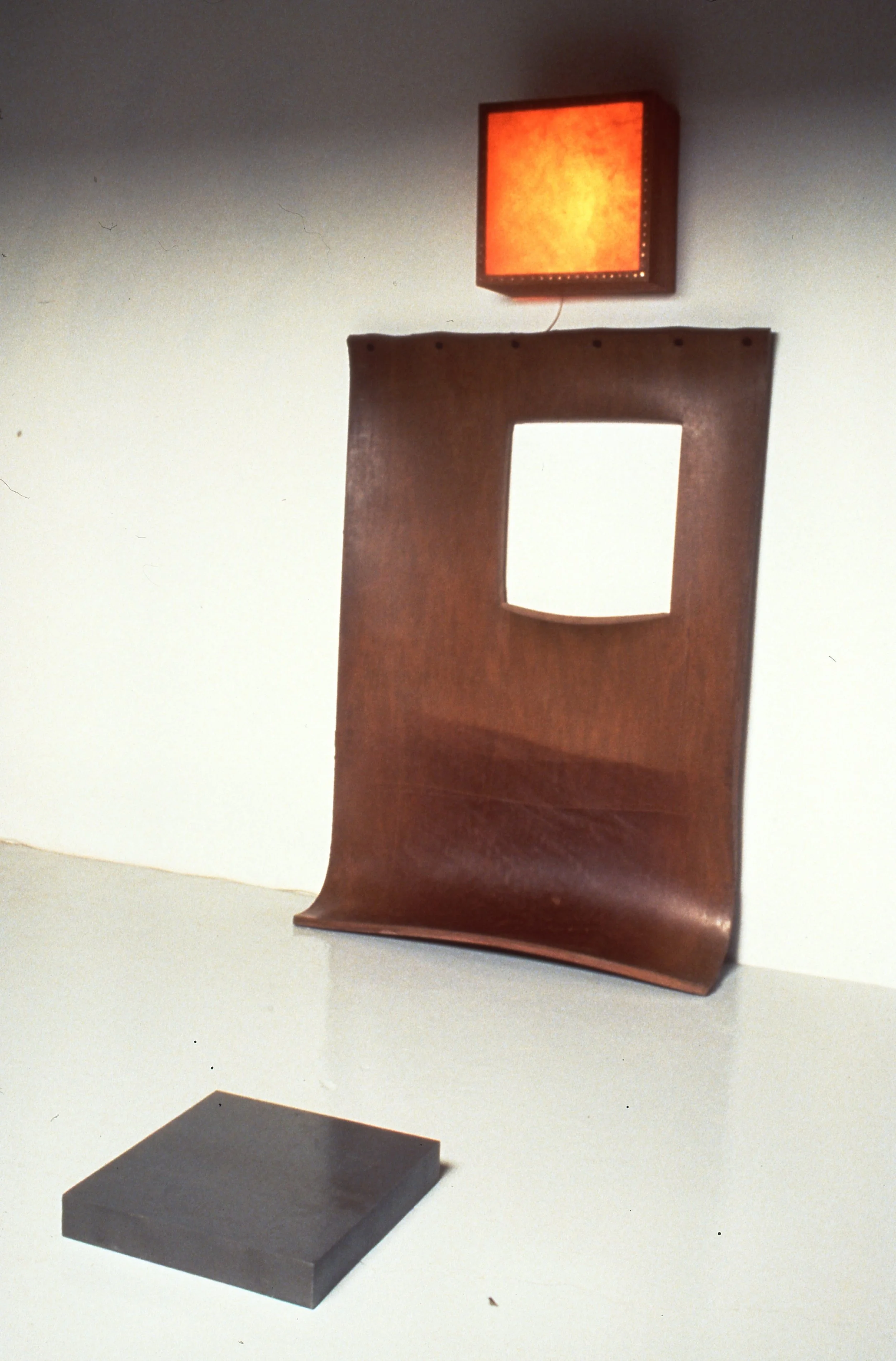 A brown wooden panel with a square cutout leaning against a white wall, a small black rectangular object on a white table in front, and a red illuminated square light fixture on the wall above.