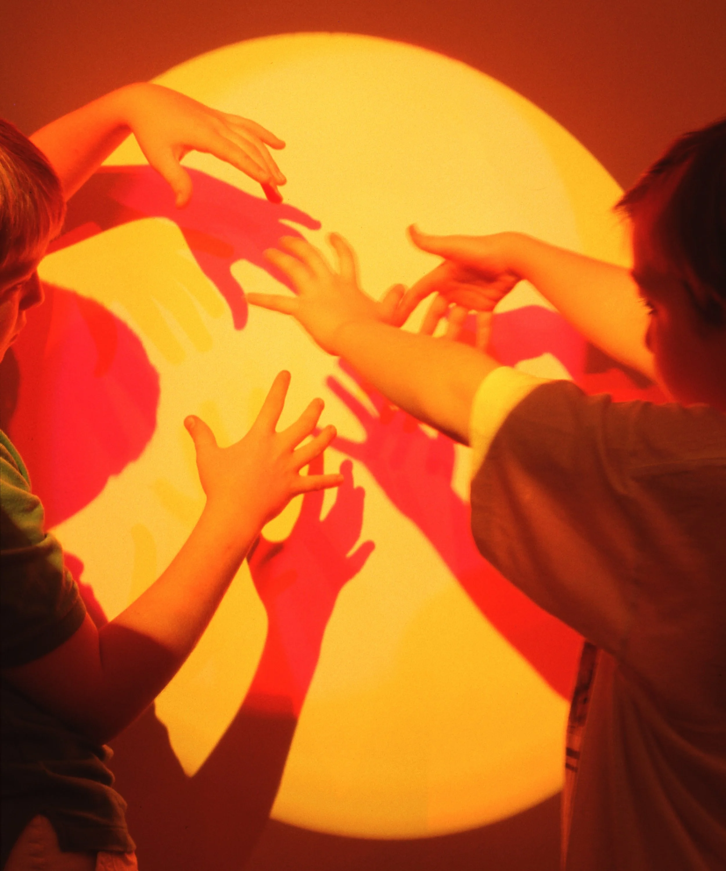 Children reaching out towards a bright, circular yellow and orange light on the wall, creating colorful shadows.