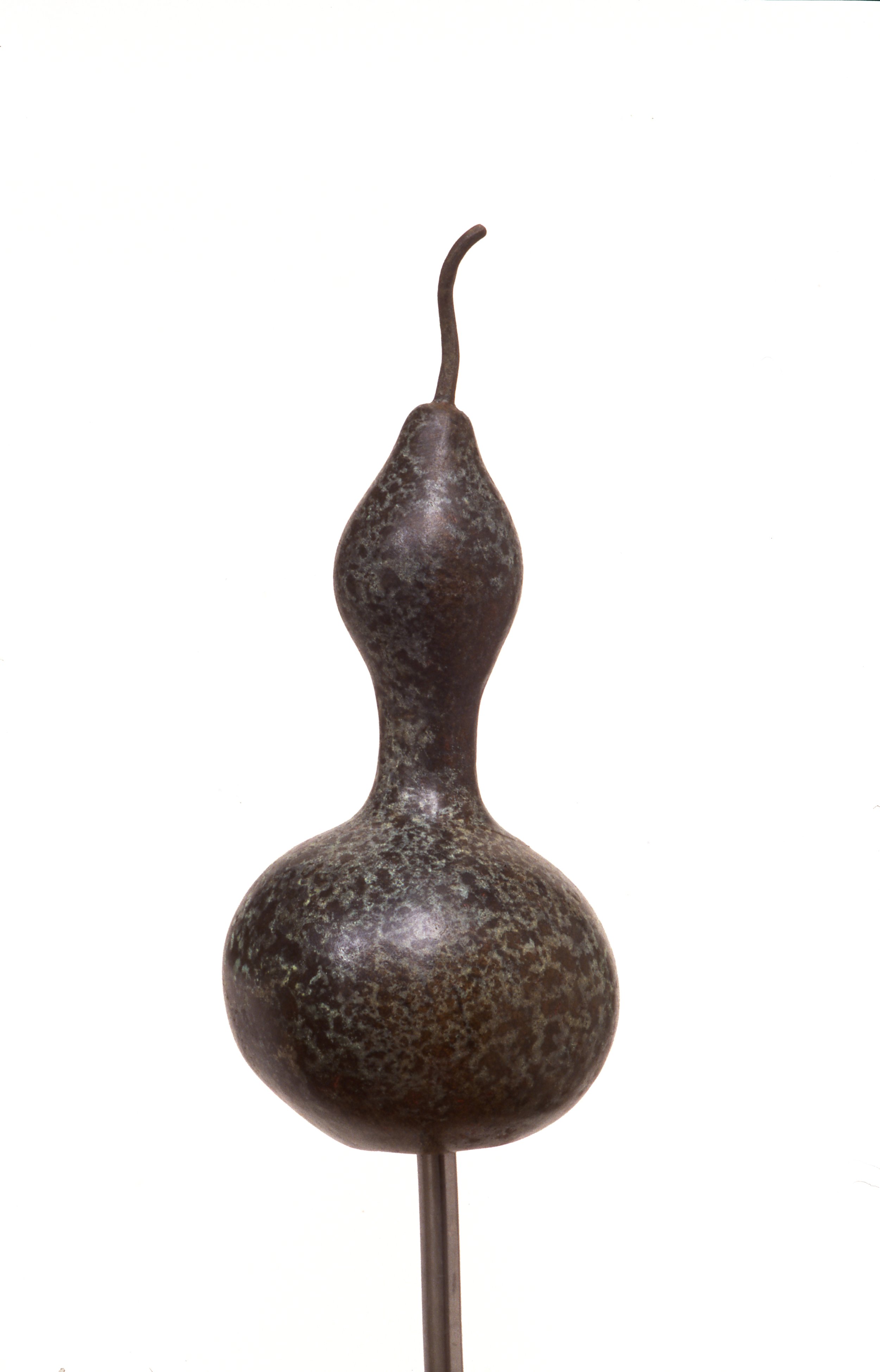 A sculptural artwork resembling a gourd with a textured, dark surface and a thin, curved stem on top, mounted on a metal stand.