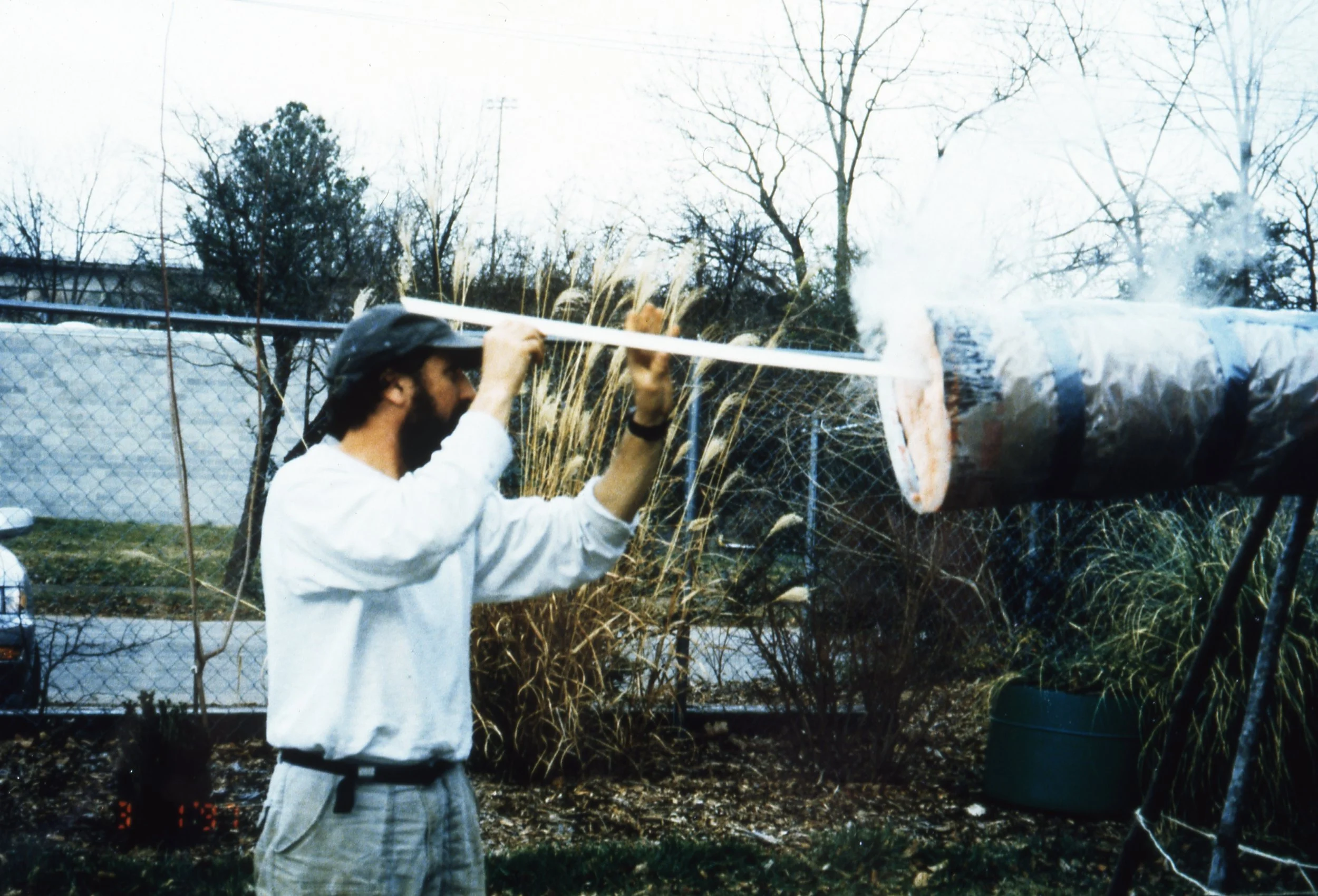 A man wearing a hat and glasses burns a paper tube with a long match outdoors, with trees and a fence in the background.