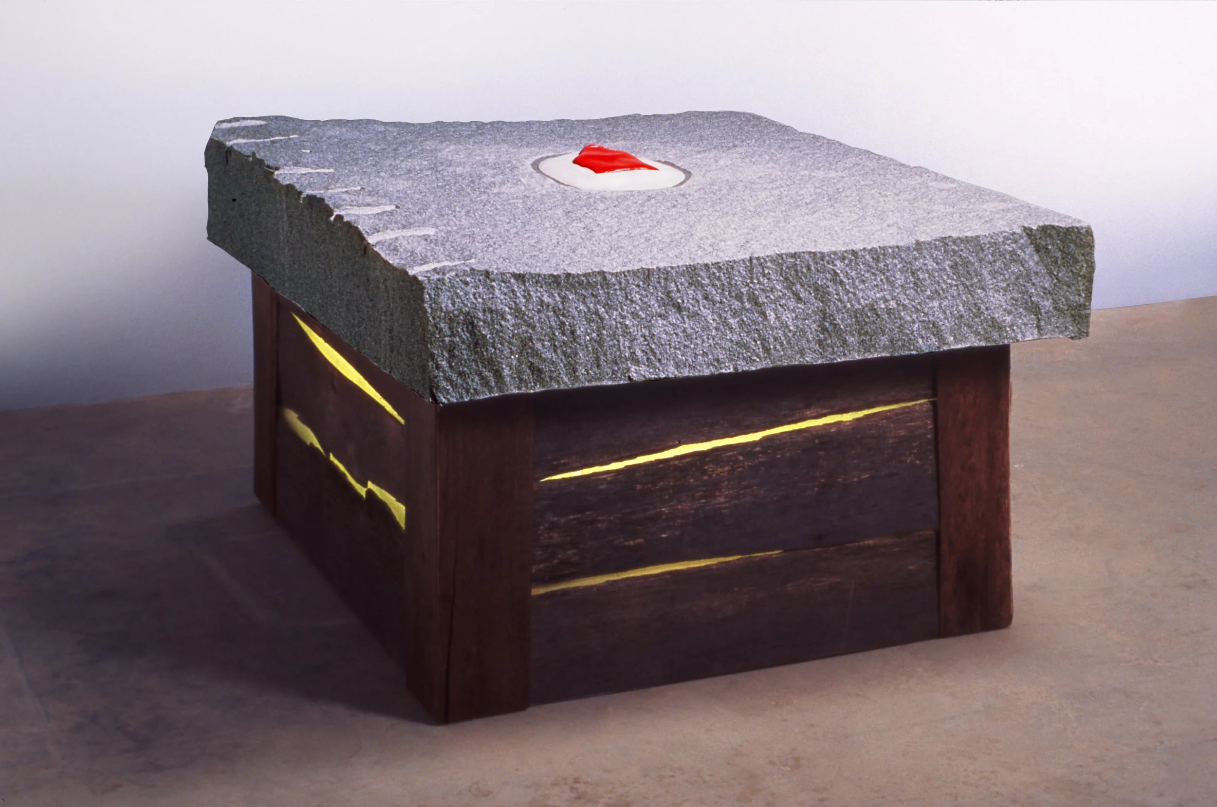 A square stone with a rough surface on top of a wooden box with yellow streaks, and a small red and white object in the center of the stone.