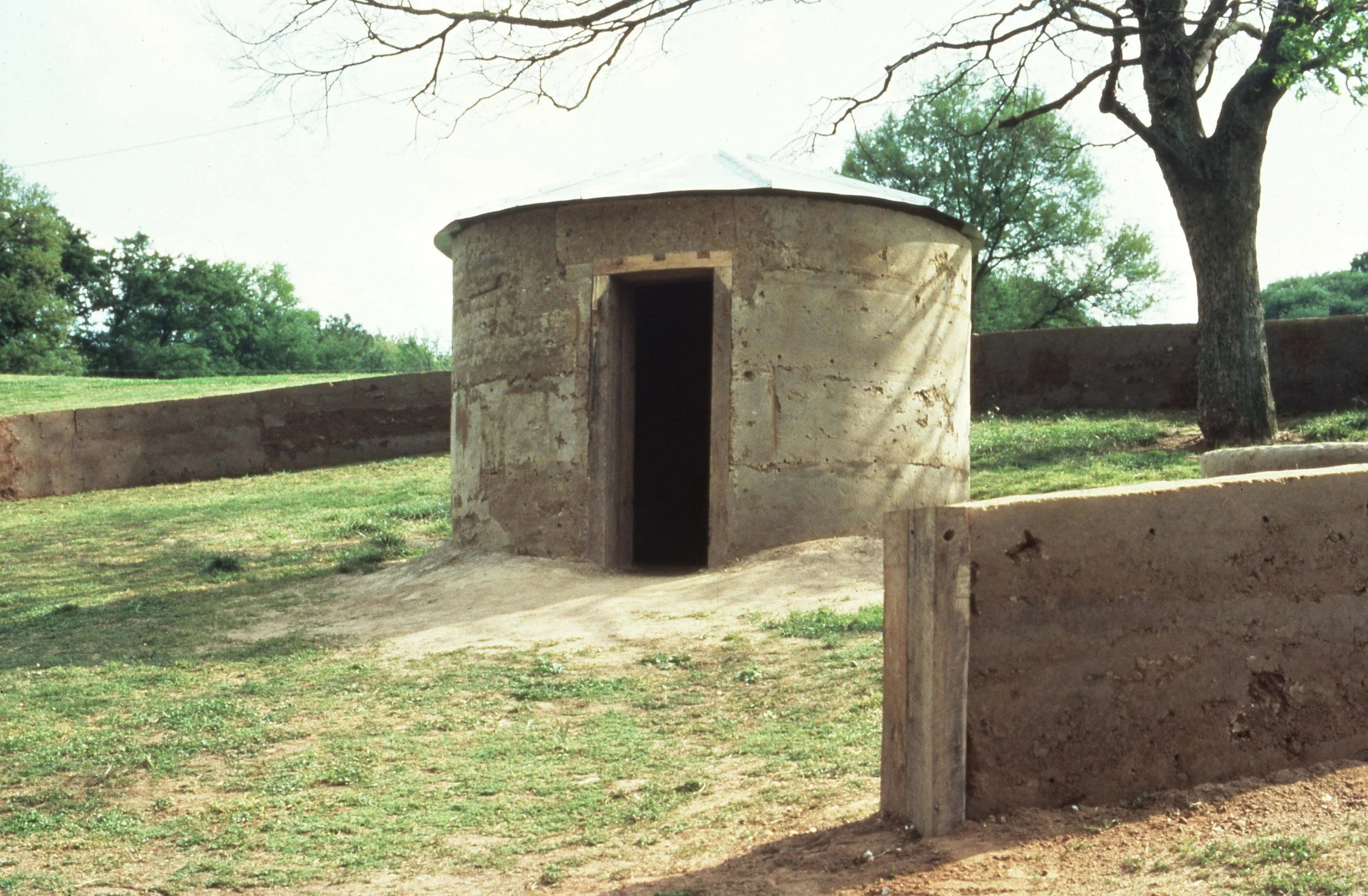 A round, stone building with an open doorway, situated outdoors on grassy terrain with trees and a low stone wall in the background.