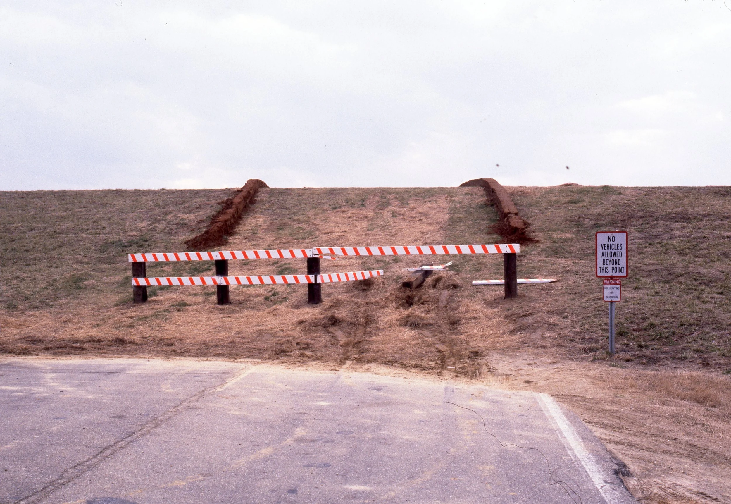 A road with a barrier and warning sign, a dirt ramp beyond the barrier, and a grassy hill in the background.