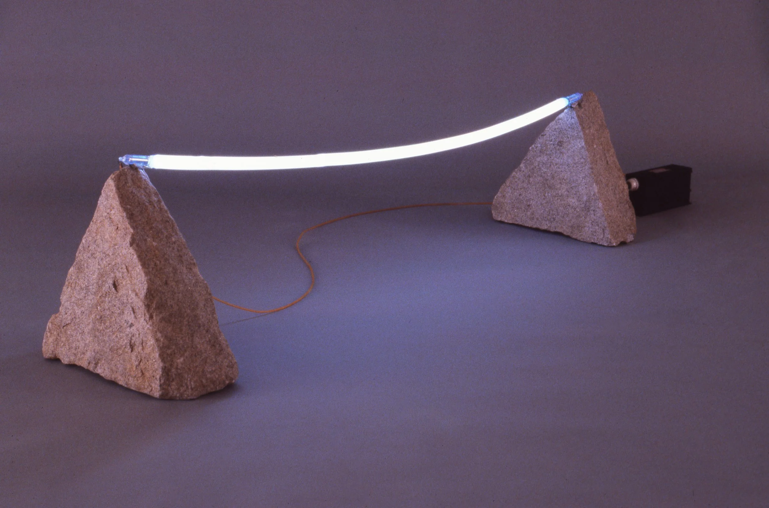 A small hoverboard made of a glowing white tube between two large rocks, with a motor attached to one rock, creating an optical illusion of a floating device.
