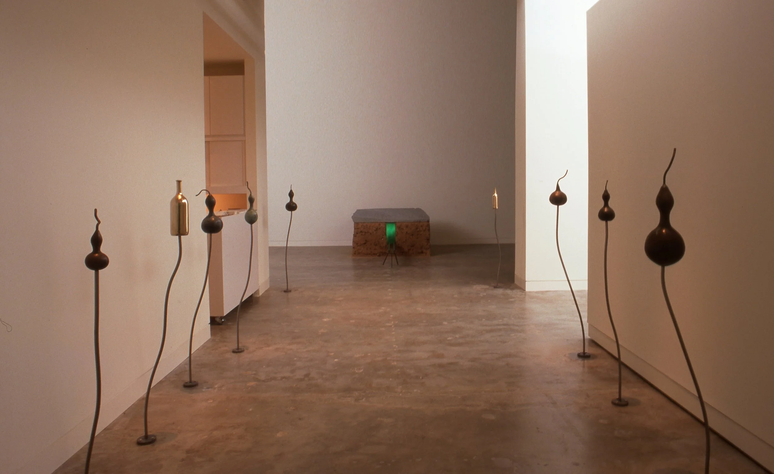 Gallery with tall, thin, abstract sculptures on curved metal stands lining the walkway, leading to a stone pedestal with a green light underneath, in a modern art exhibit.