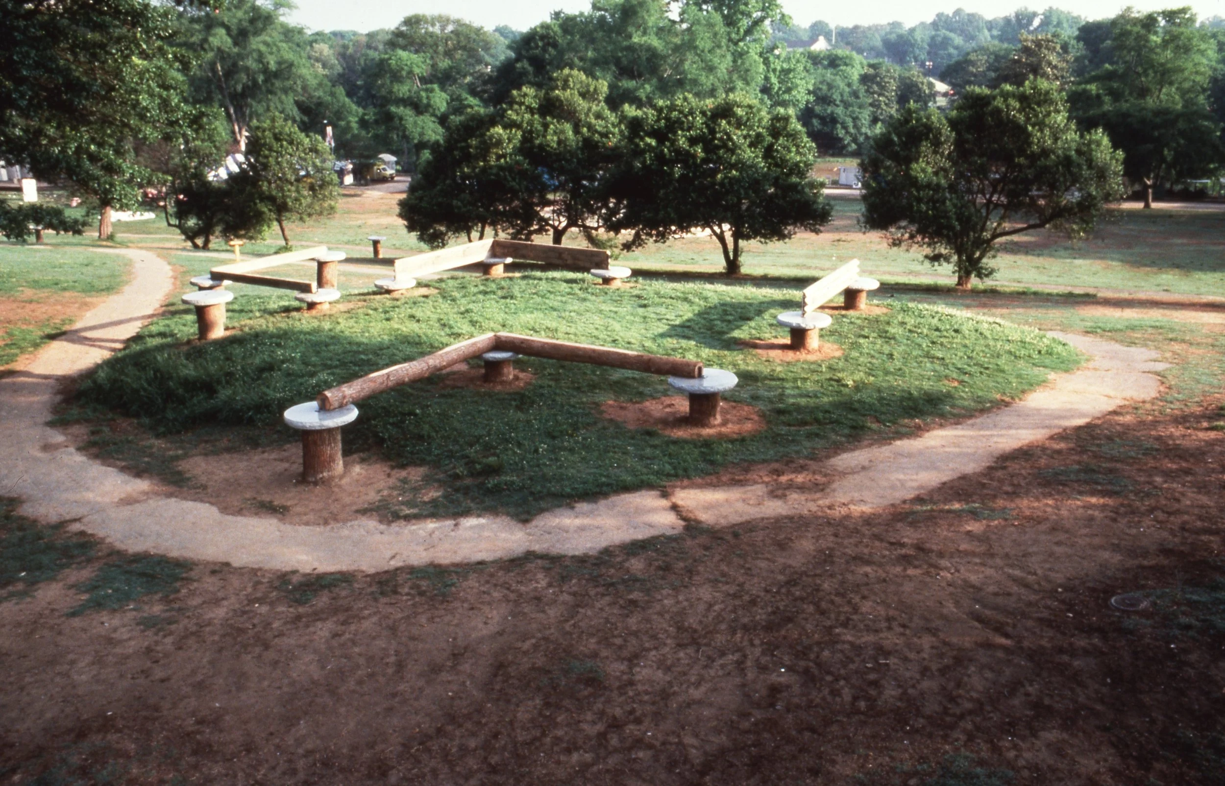 A park with a circle of wooden balance beams on concrete supports, surrounded by grass and dirt pathways, with trees in the background.