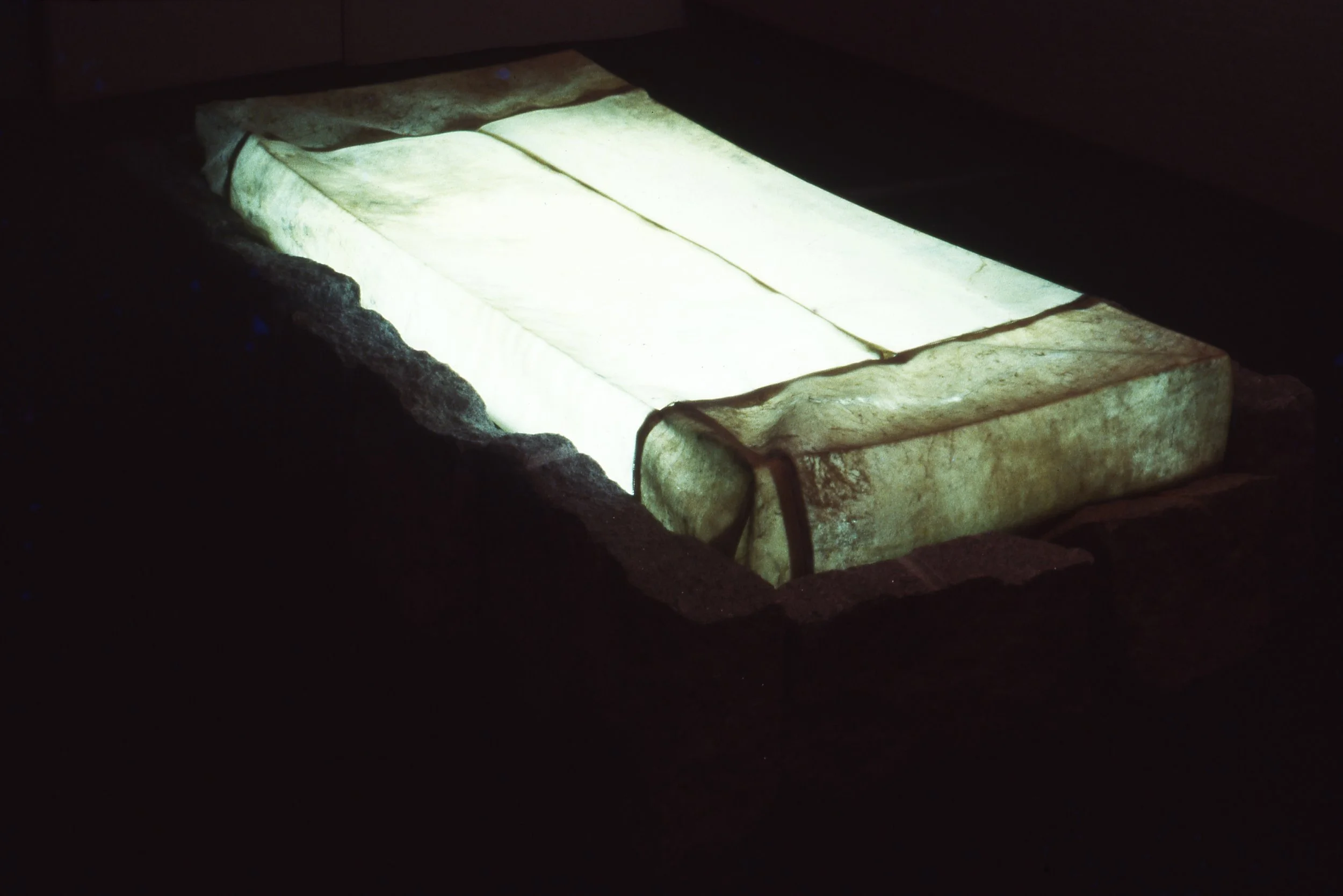 A rectangular sink or basin carved from a single piece of light-colored stone, illuminated from above, with rough edges and corners, set into a dark surface or structure.