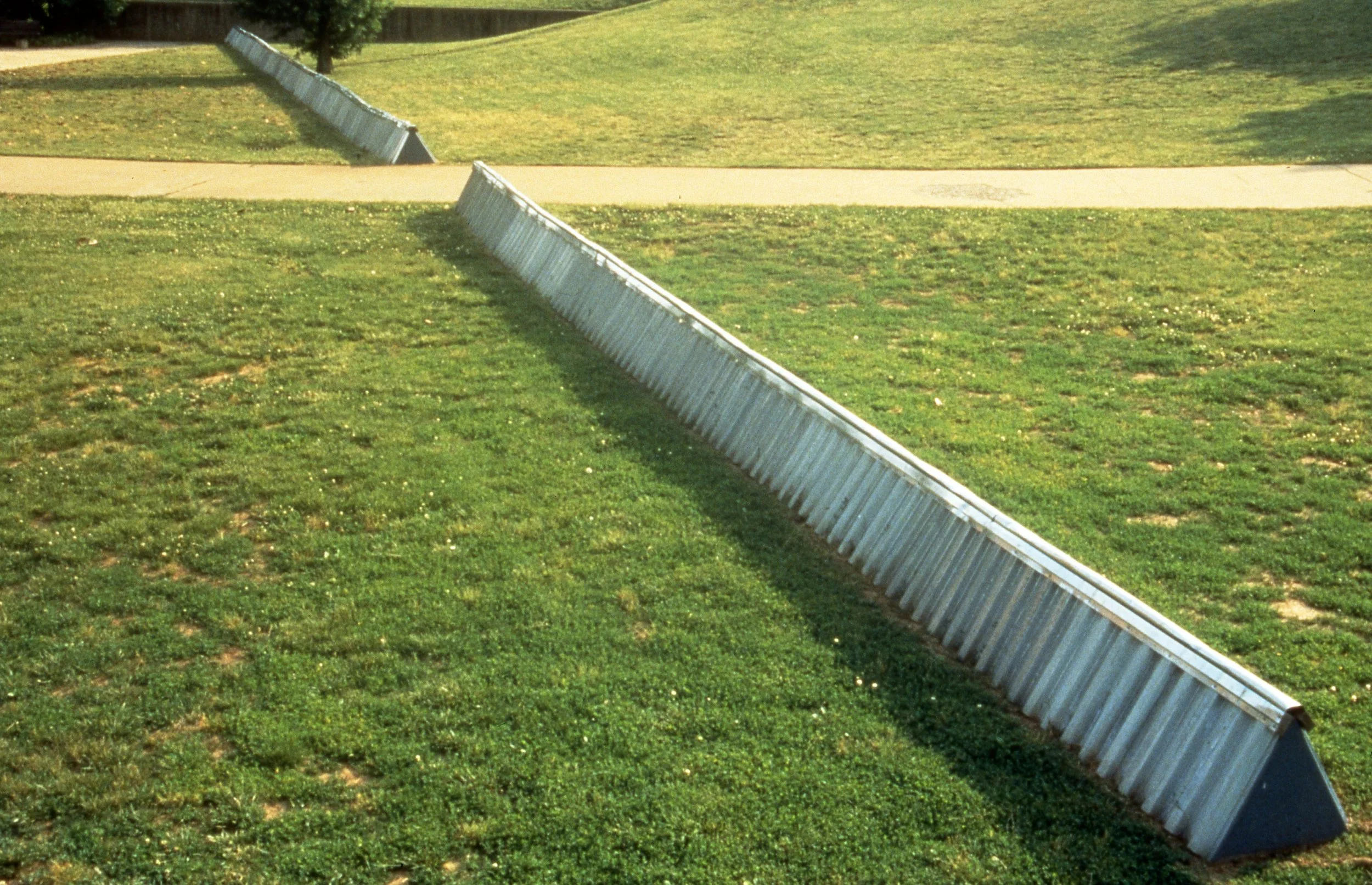 Ski jump ramp on a grassy field near a sidewalk with a tree in the background.