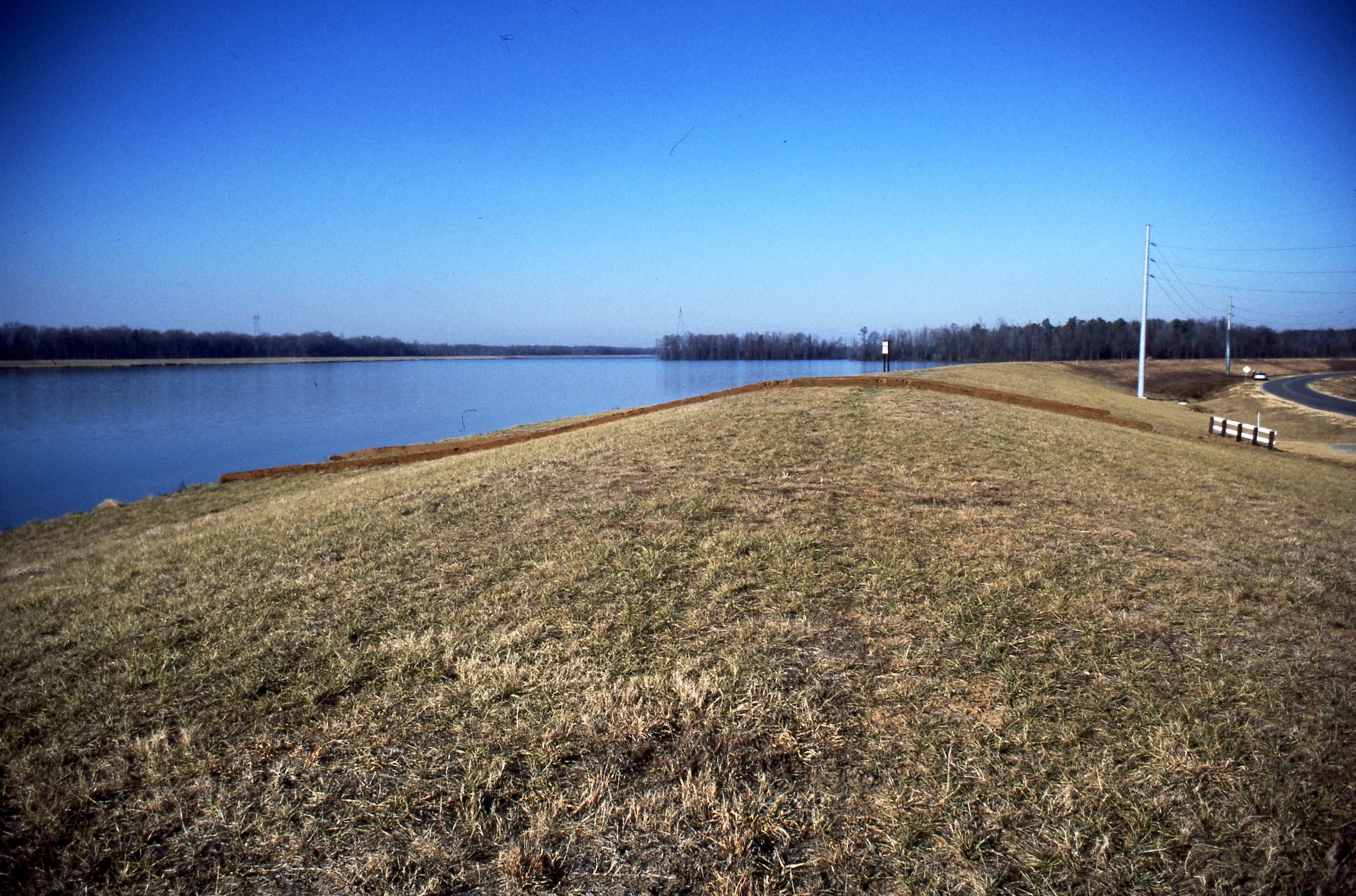 A grassy hillside by a calm river with a clear blue sky, power lines, and a curved road on the right side.