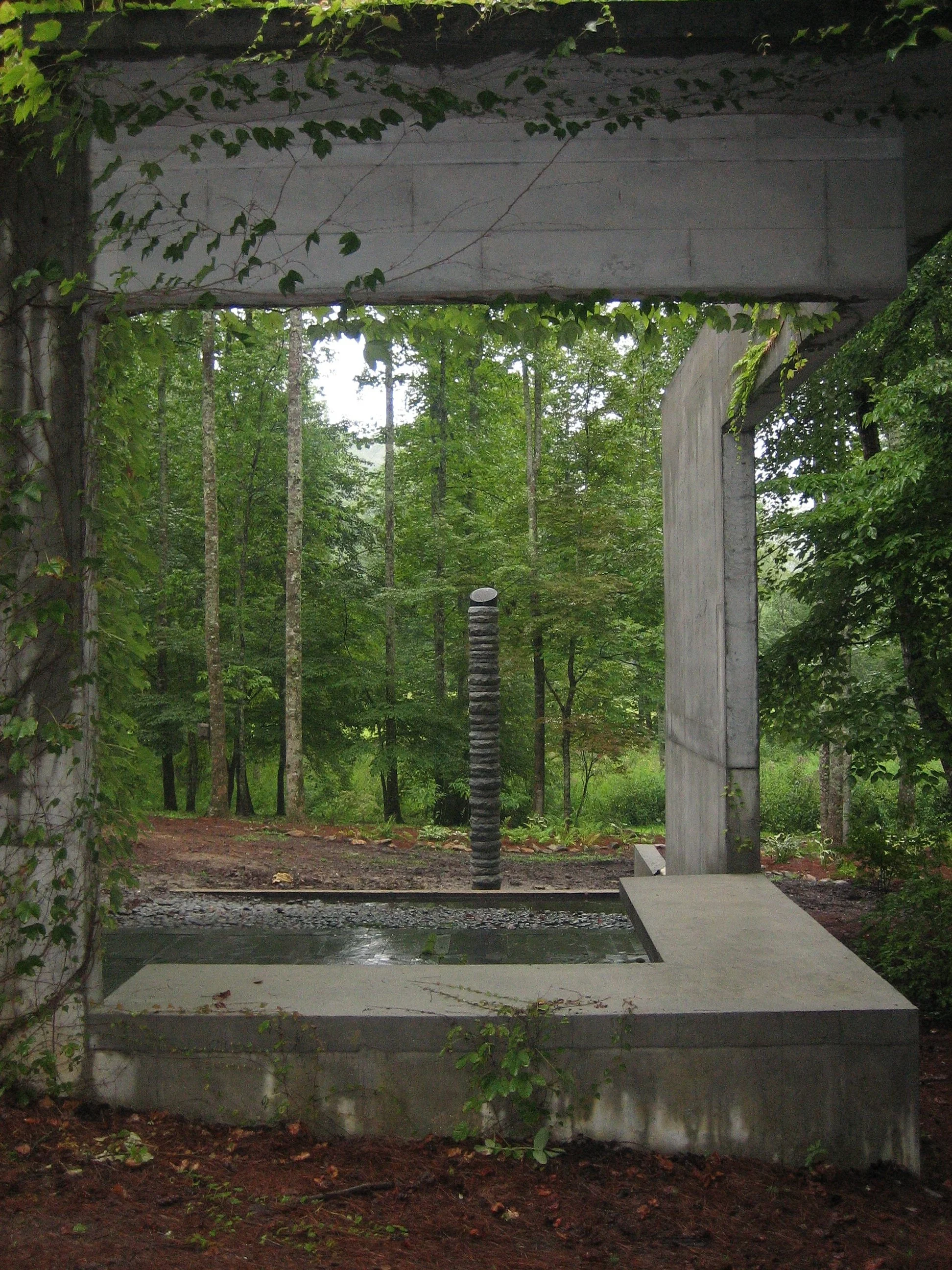 Modern outdoor water fountain with a vertical stack of flat, round stones surrounded by trees and foliage.