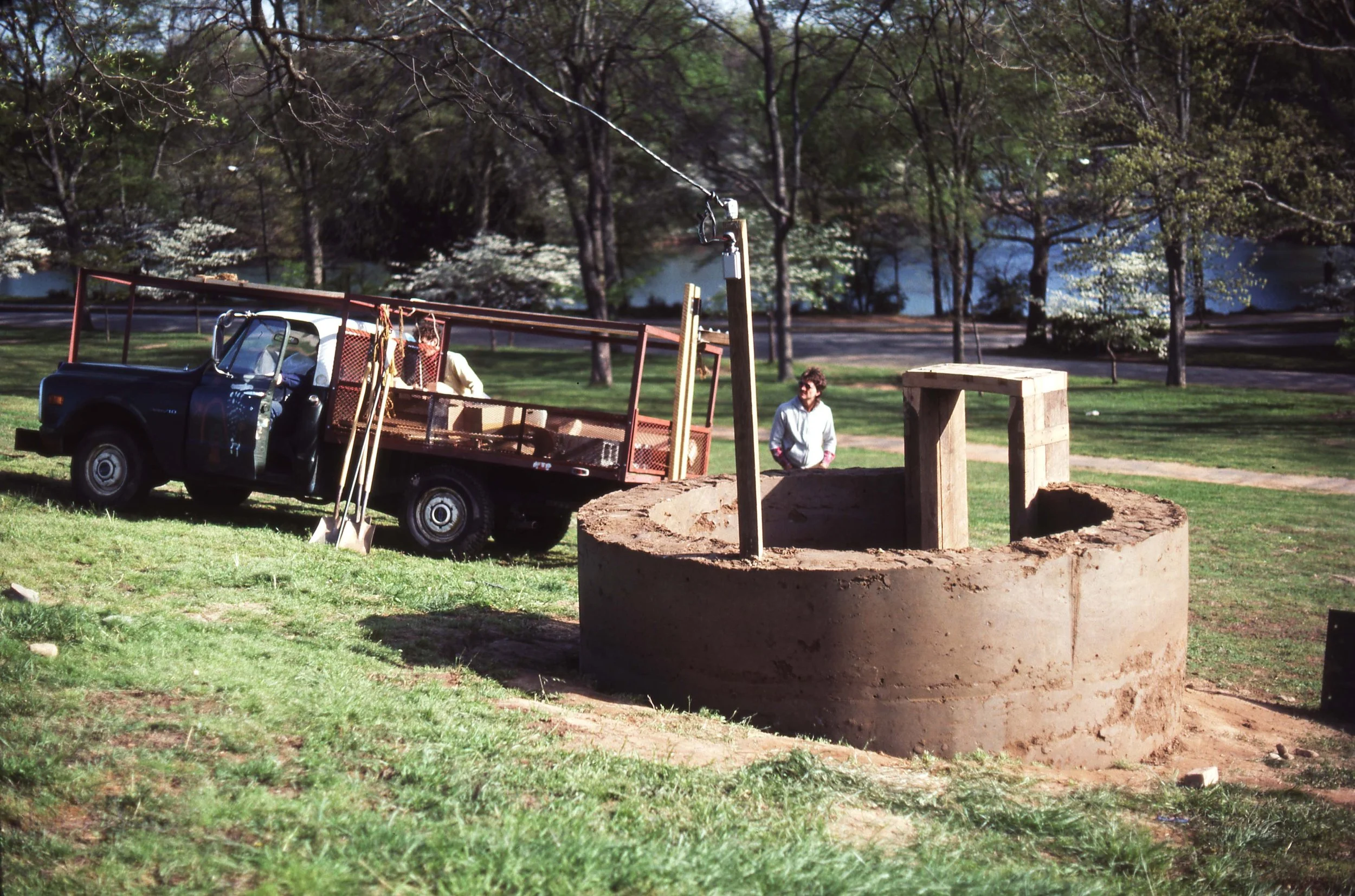 A man working on construction of a well or circular structure in a park with grass, trees, a river, and a road in the background.