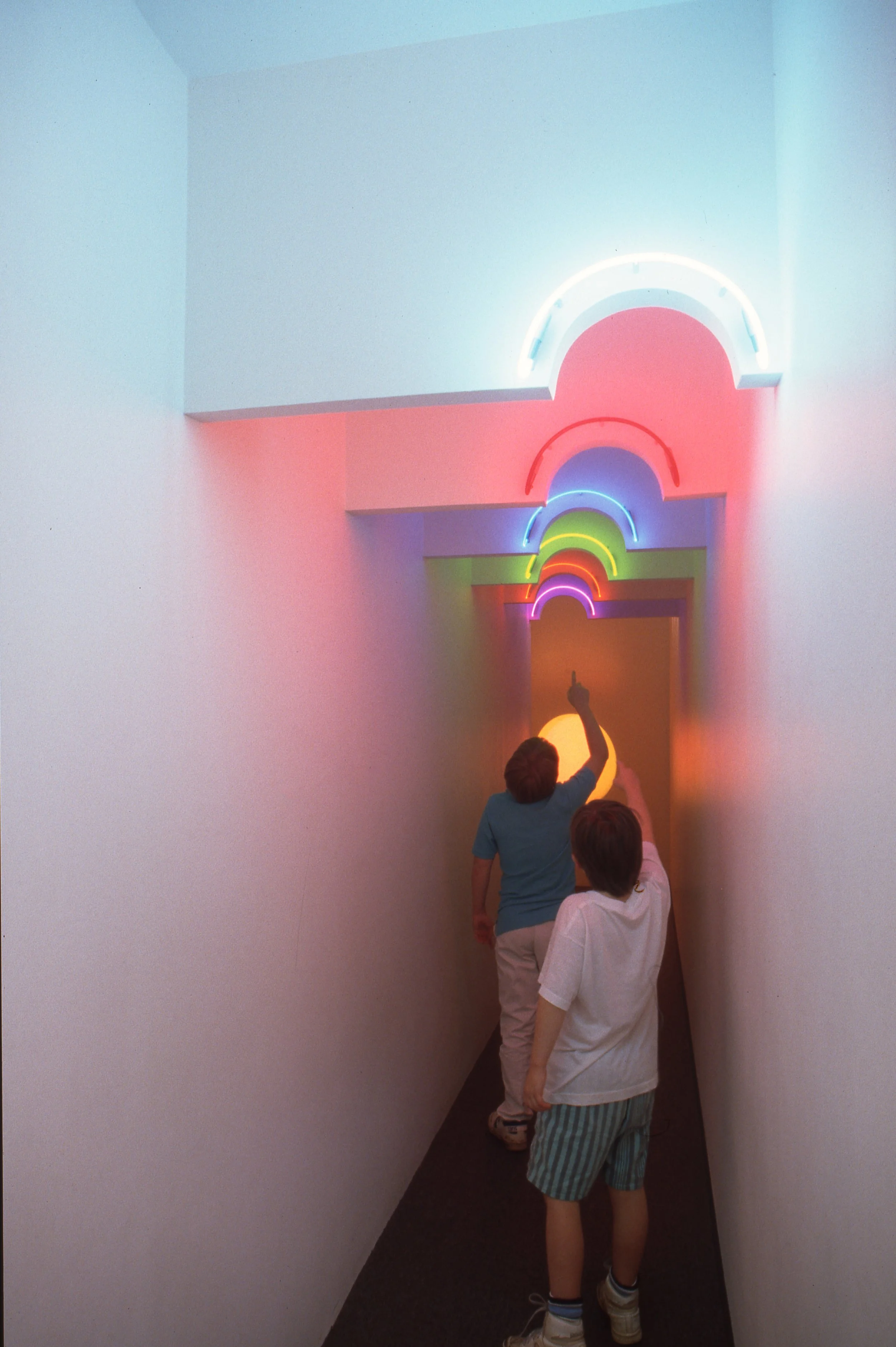 Two children walking through a narrow hallway with colorful neon rainbow arch decorations on the ceiling, with one touching the wall.
