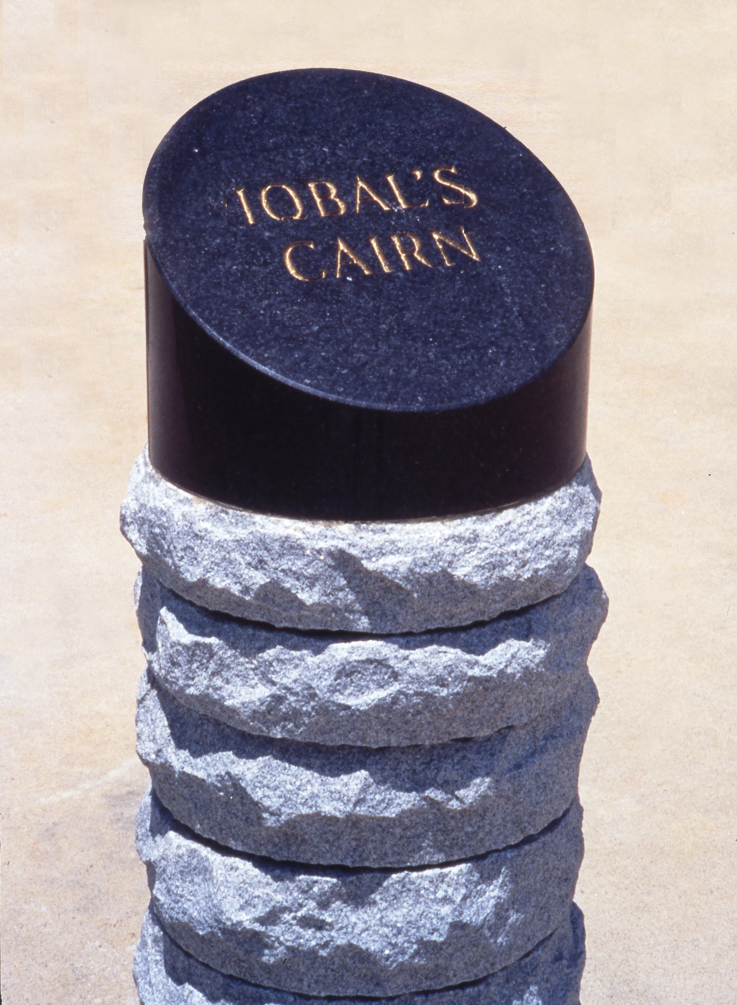 A black cylindrical stone with the words "IOBAL'S CAIRN" engraved in gold on top, placed on a stack of five rough, light-colored rocks.