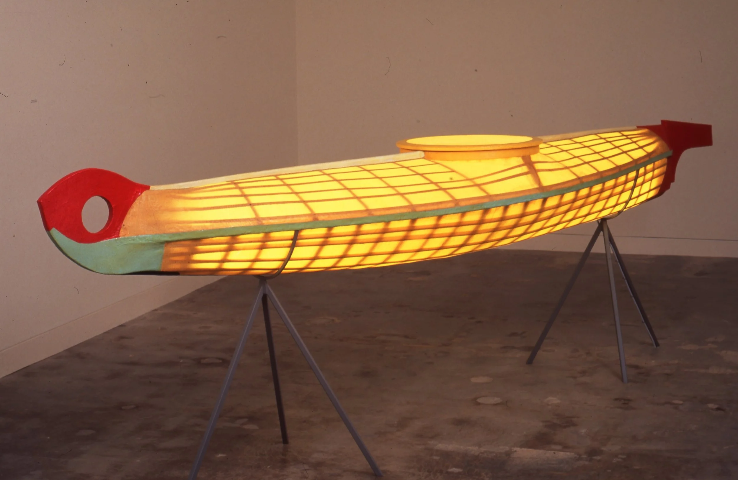 Illuminated wooden boat sculpture on black metal stands in an indoor gallery setting.