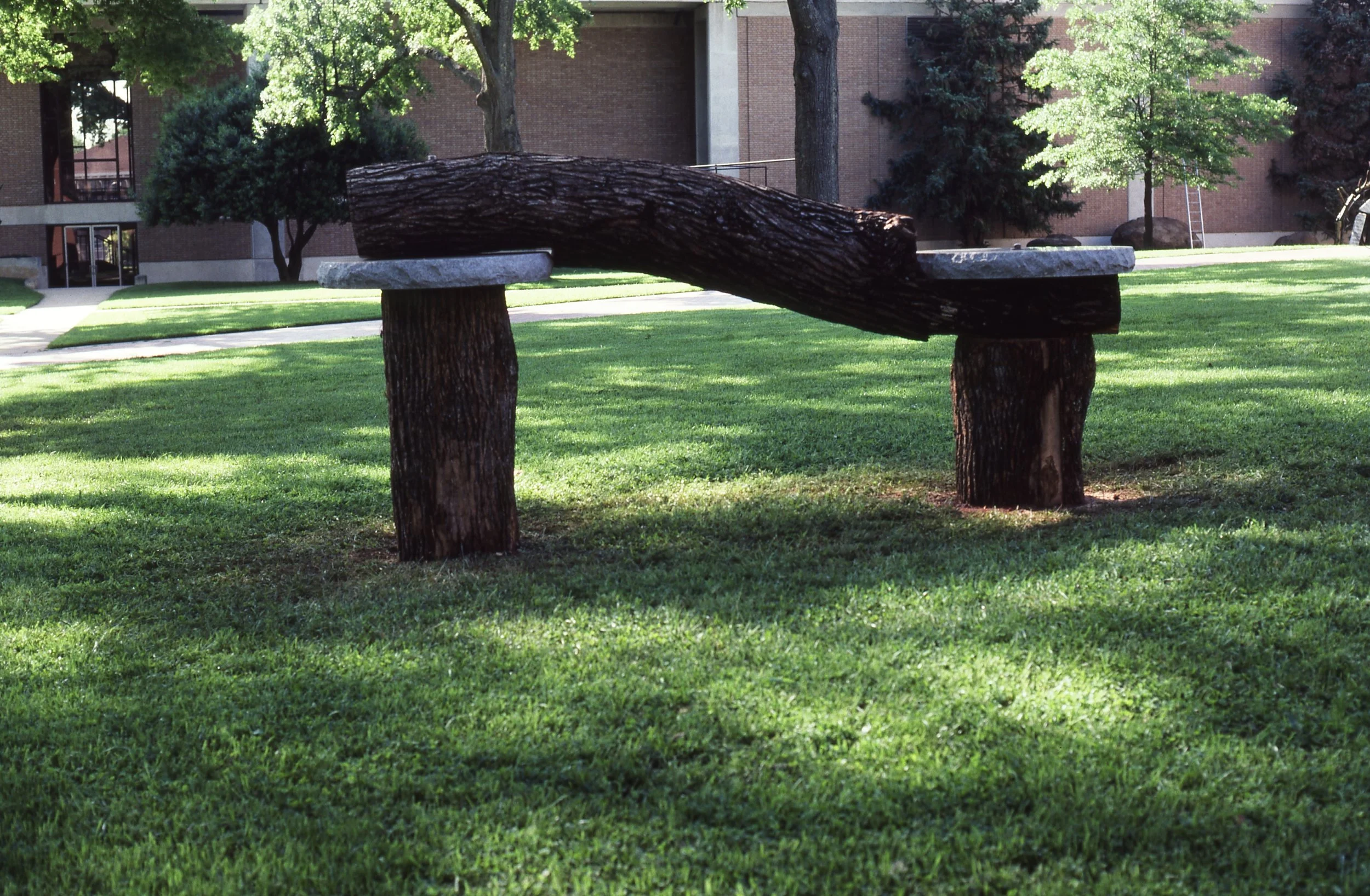 A park bench made from two large tree stumps and a thick log in a grassy area with trees and a brick building in the background.