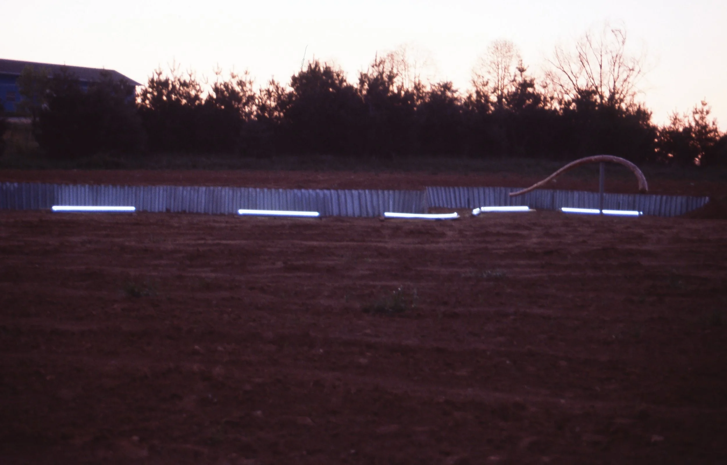 Construction site at dusk with a curved pipe, metal fencing, and illuminated lights on the ground.