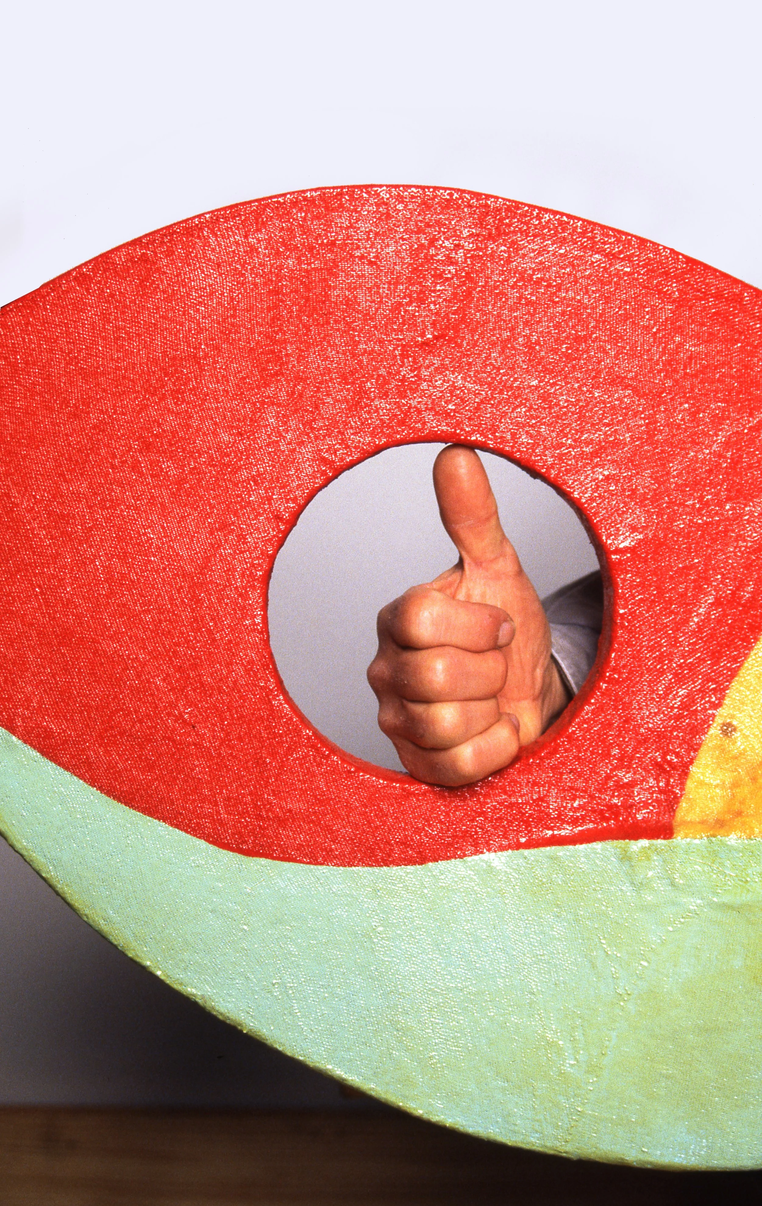 Person giving a thumbs-up behind a large, textured, colorful prop resembling a slice of watermelon.