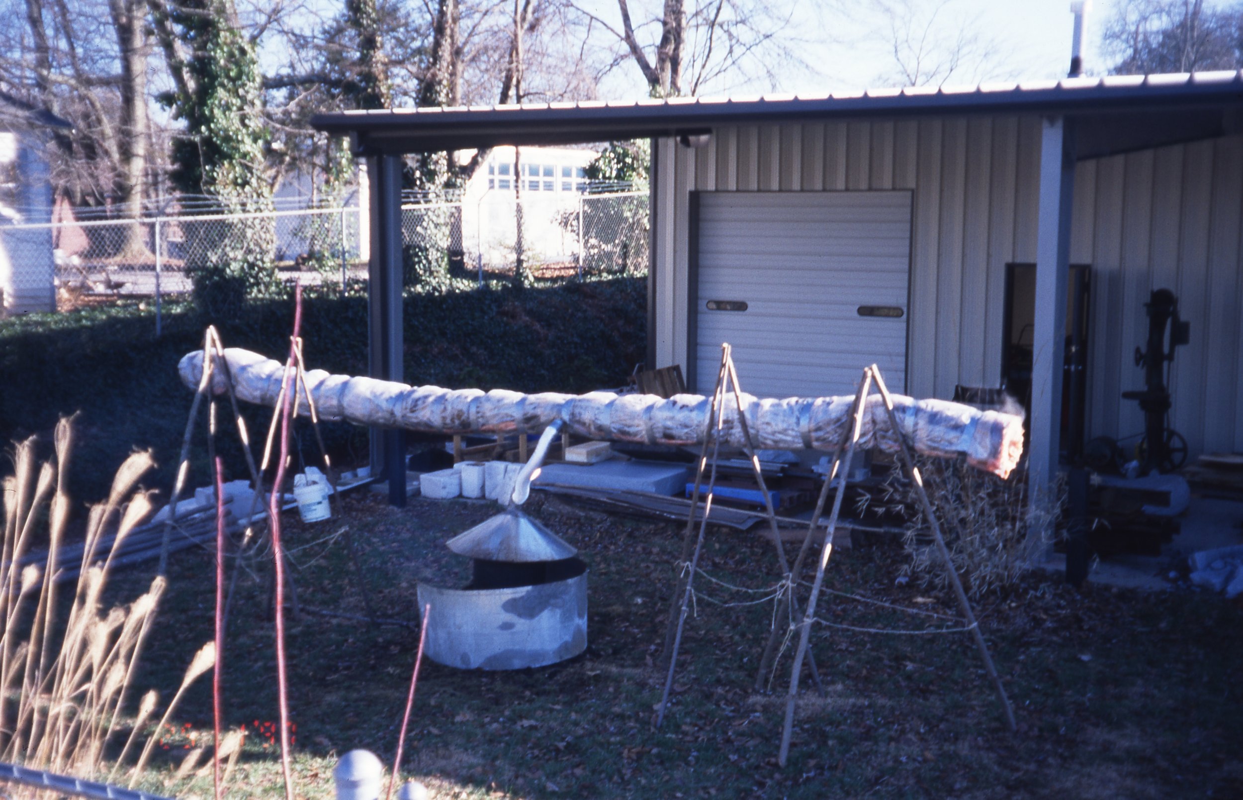 An outdoor scene showing a large log supported by wooden tripods in front of a small building with a roller door and a side door. There are woodworking tools and supplies nearby, including a metal fire pit in the foreground, and trees and a fence in 
