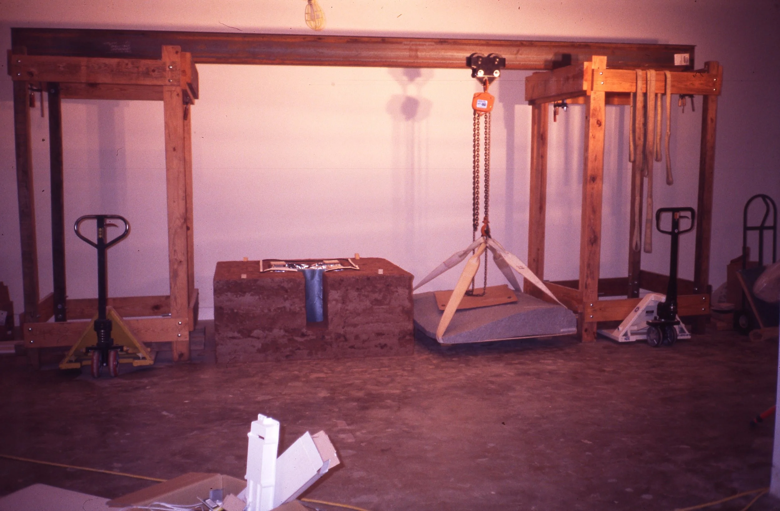 A workshop with wooden frames, a hydraulic jack, a chain hoist, and a pallet jack, with tools hanging from the frames and miscellaneous items on the floor.