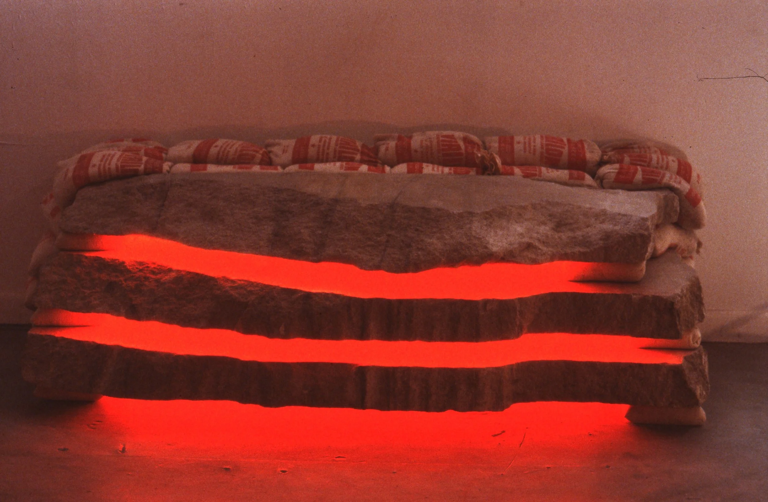 A piece of furniture made of stacked, unevenly cut slabs of stone or concrete, illuminated from below with red lighting, placed on a plain floor with a blank wall in the background.