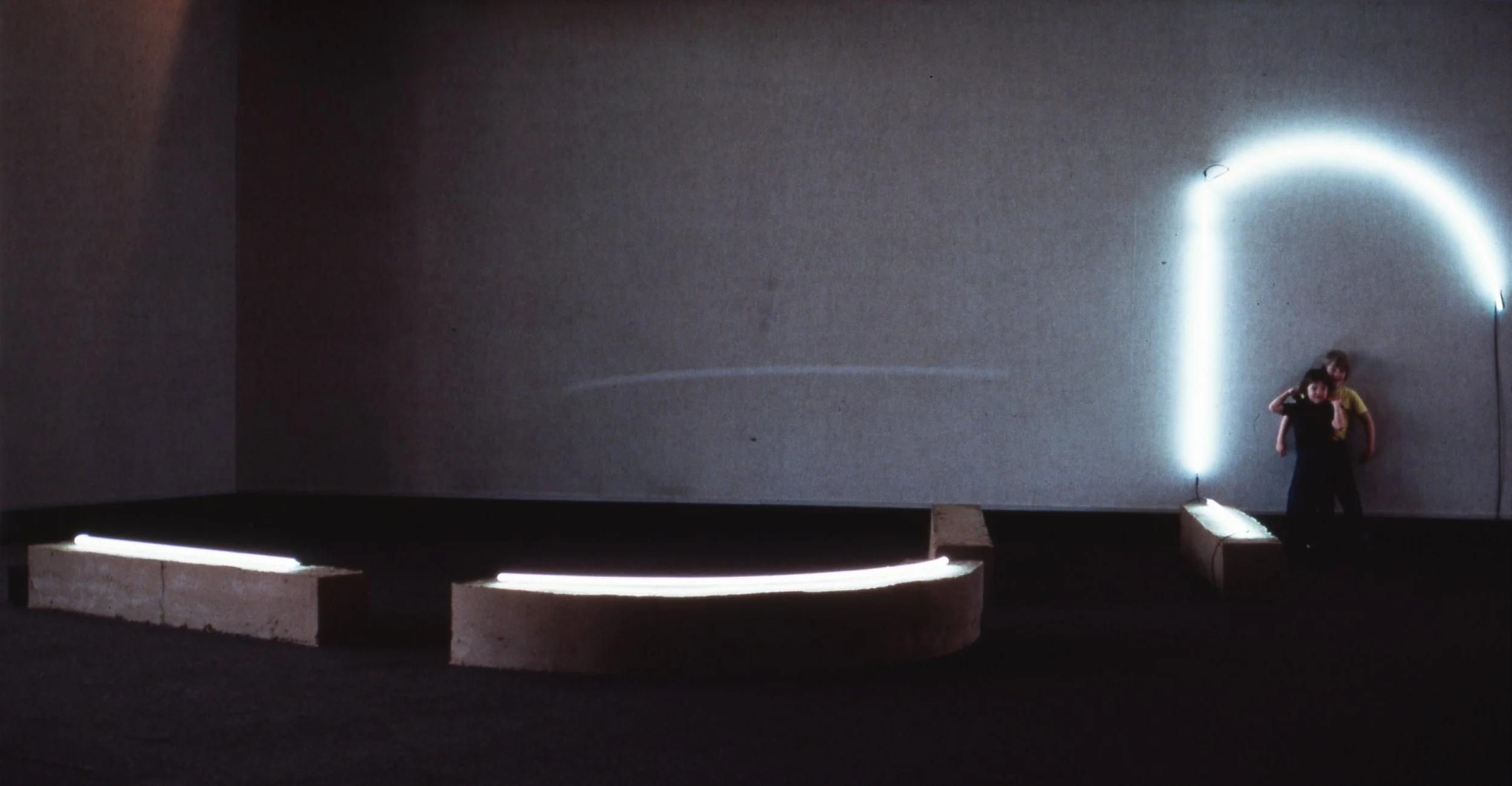 Two children stand under a glowing, curved neon light on a plain, gray wall inside a dark room.