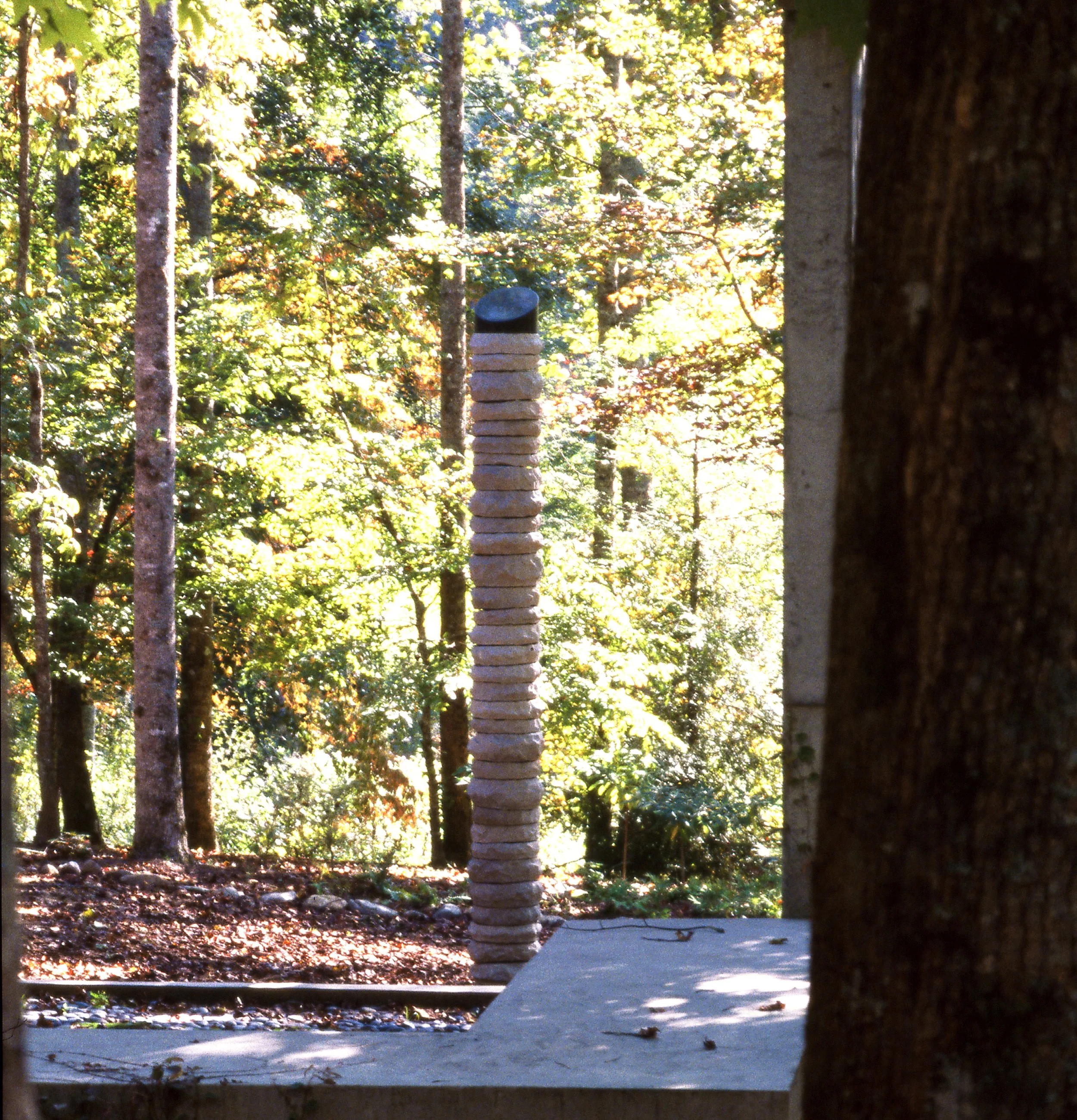 A stack of flat stones arranged on a concrete platform in a forest setting with trees and sunlight in the background.