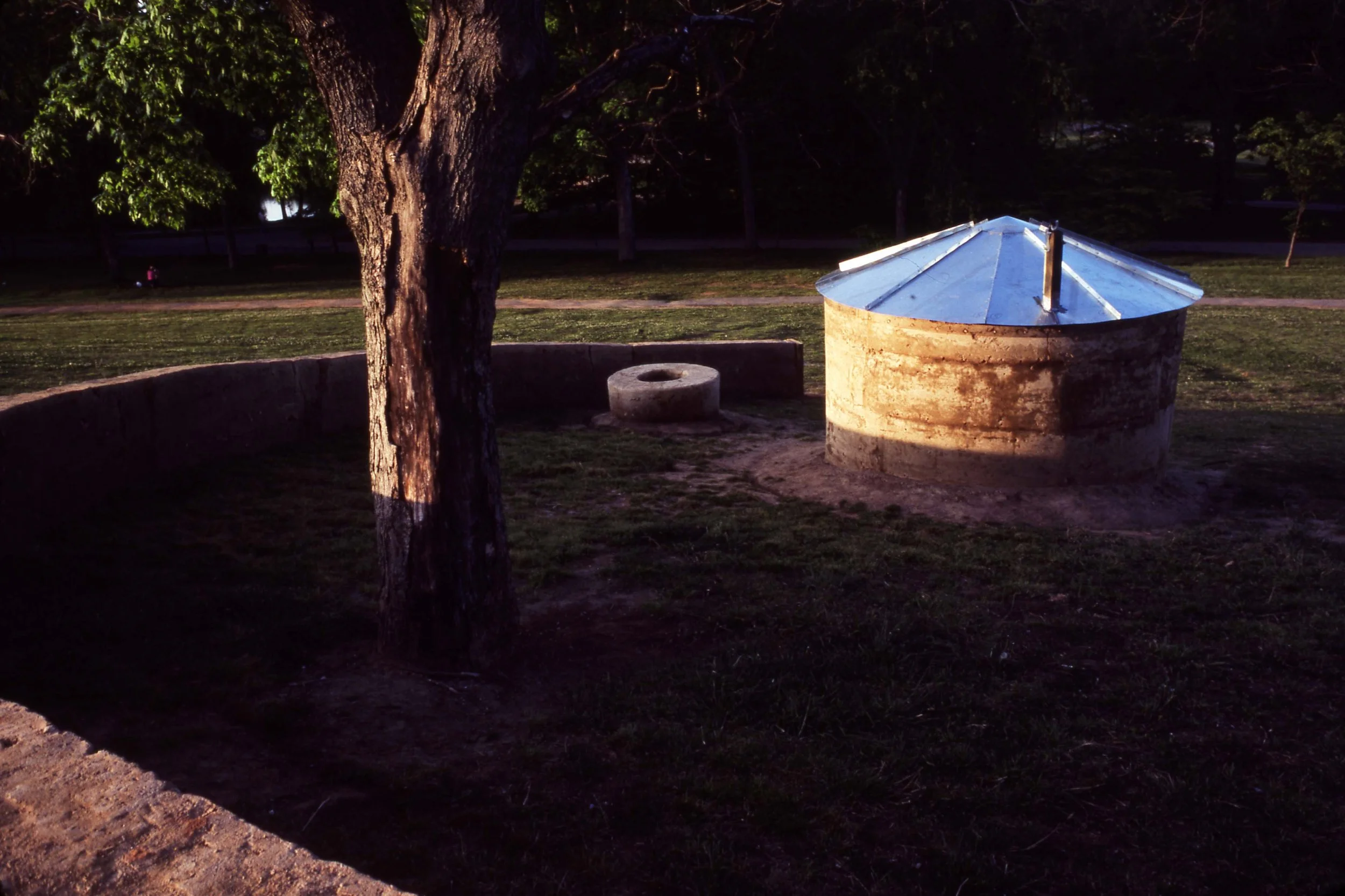 A park scene at night with a large tree, a circular concrete well, and a small round concrete structure with a hole on top, illuminated by a warm light.