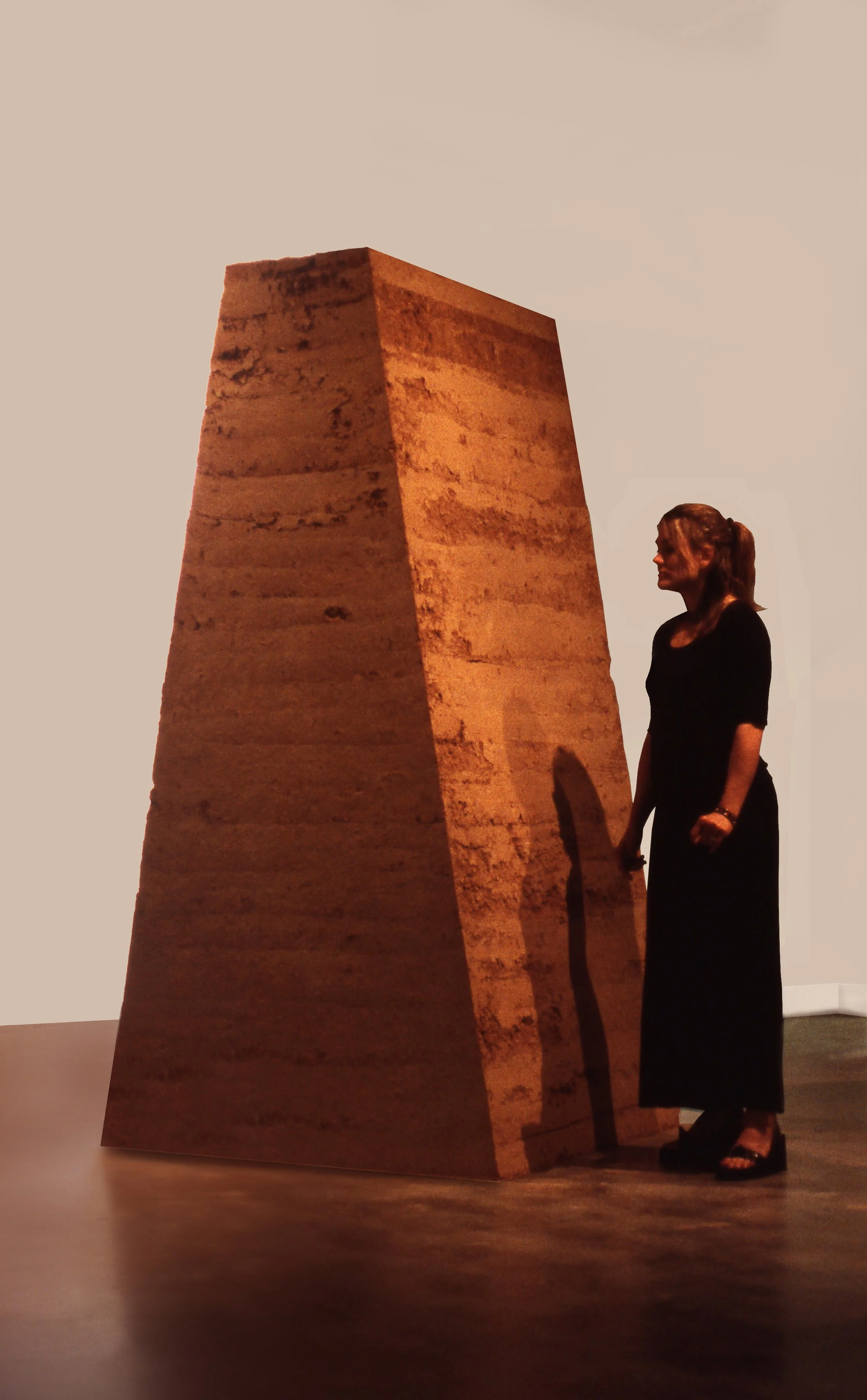 A woman in black dress standing next to a tall, rectangular, stone structure with a textured surface, inside a gallery or museum.
